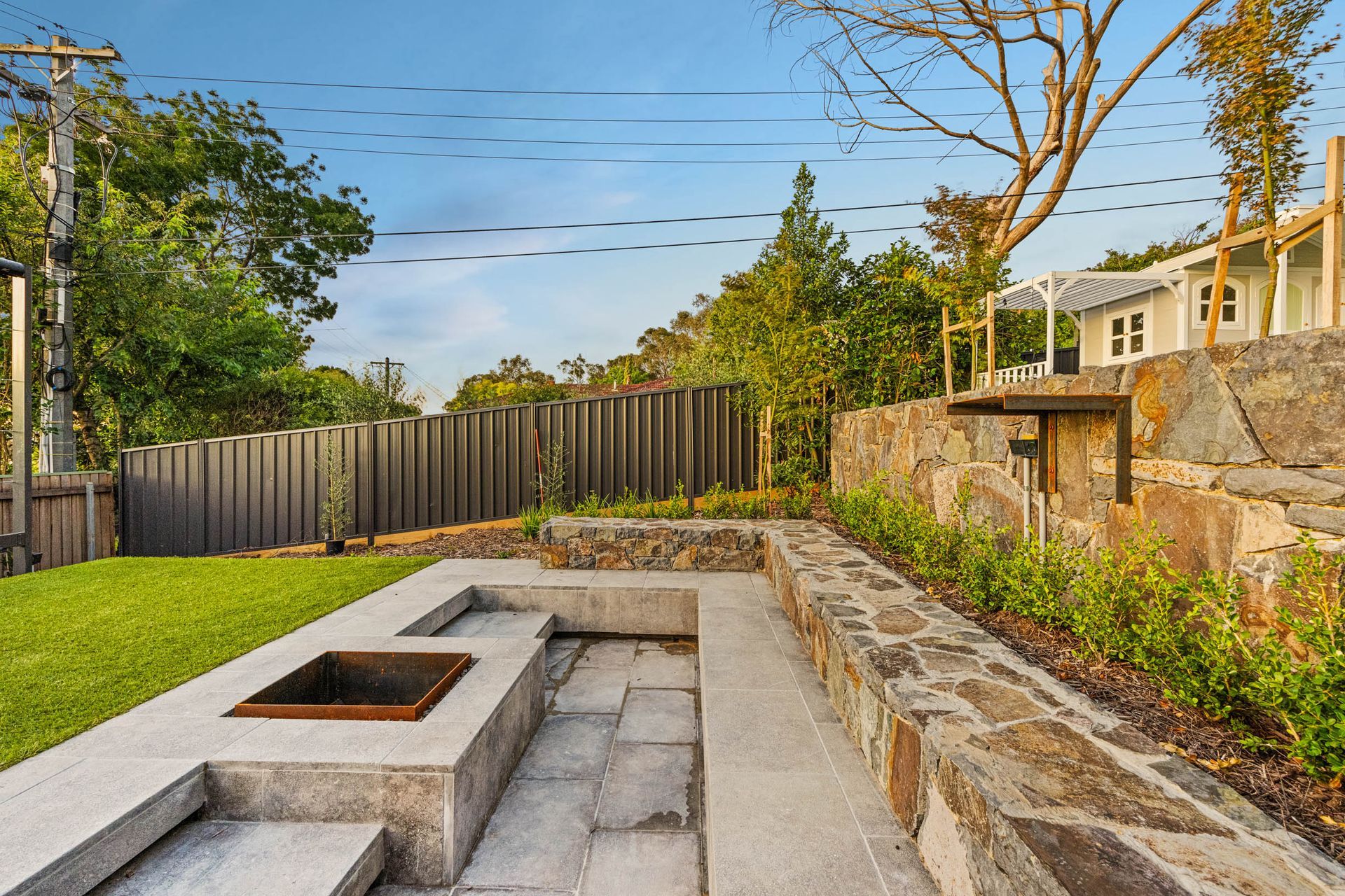 A sunken stone fire pit patio built into a terraced garden with a natural stone retaining wall and grassy lawn.