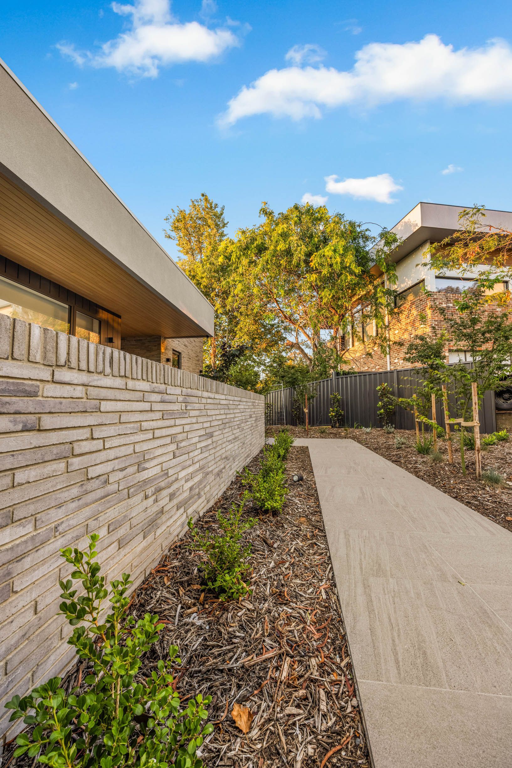 A straight concrete path runs alongside a long, textured light-grey brick wall, bordered by mulch and small shrubs.