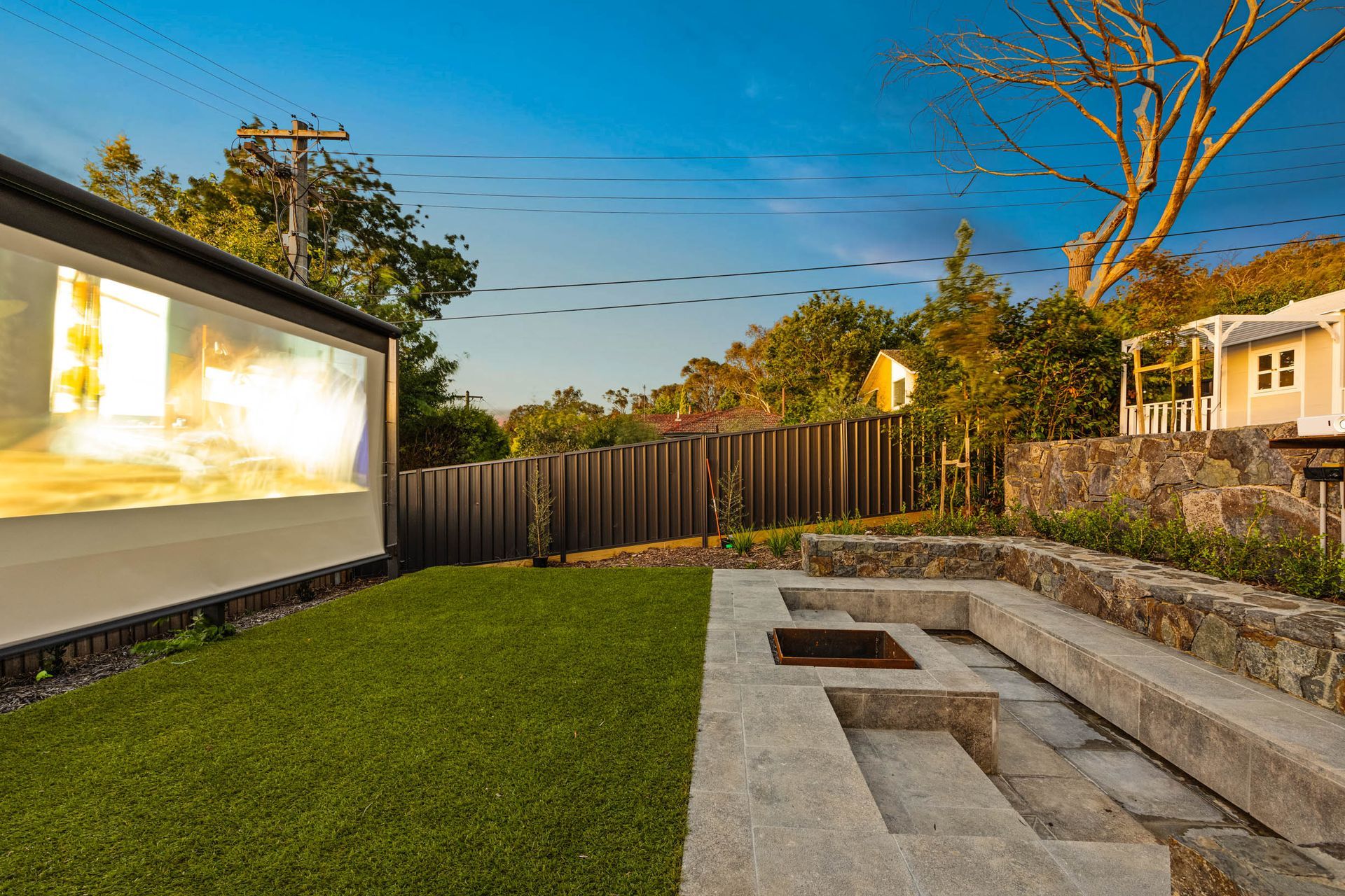 A sunlit backyard featuring a stone fire pit with built-in bench seating, artificial grass, and a large projection screen.