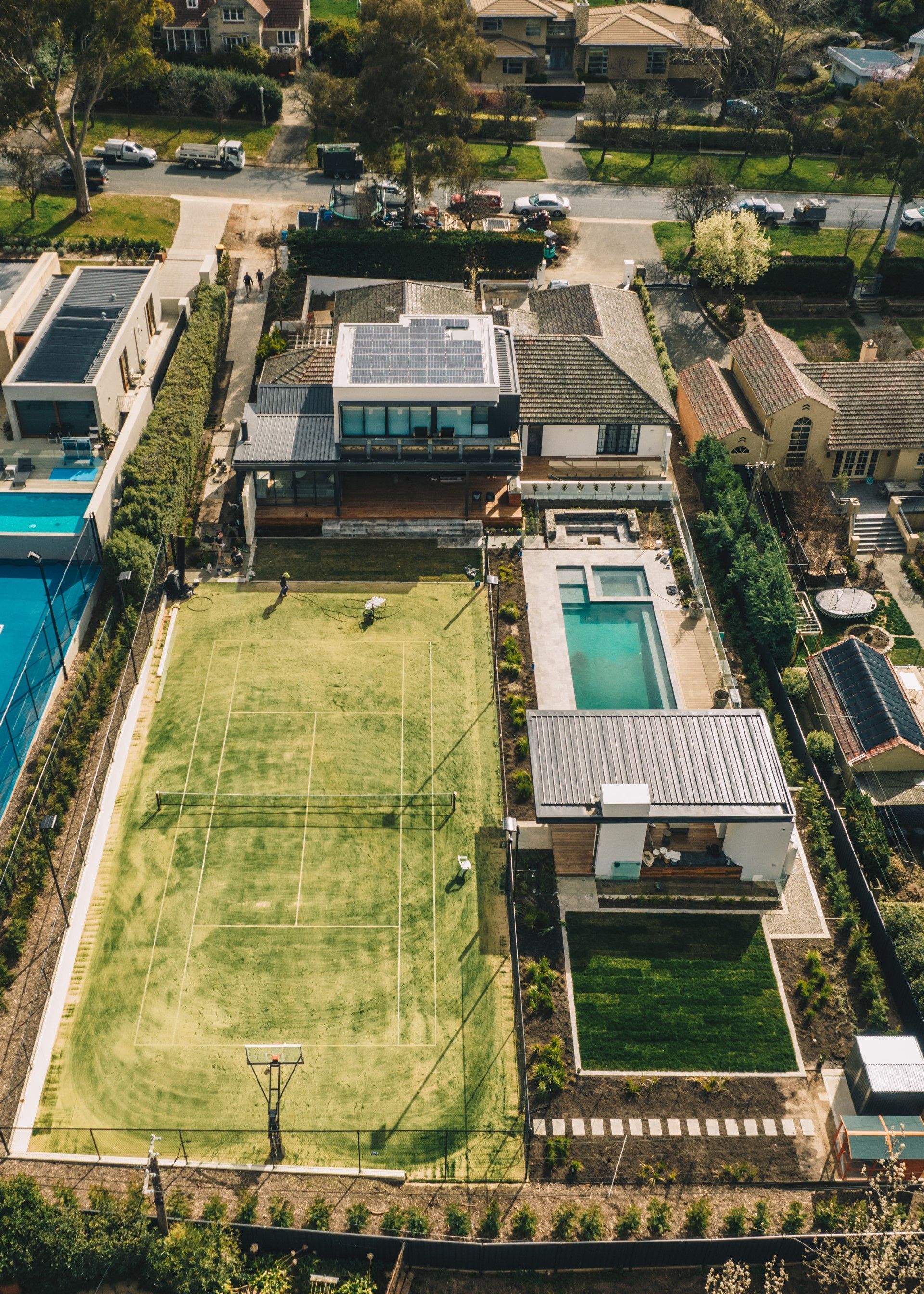 An aerial view of a luxury residential backyard featuring a grass tennis court, a swimming pool, and a modern home.