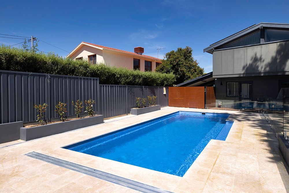 A rectangular swimming pool with blue water, surrounded by light-colored stone pavers, gray fencing, and a modern house.