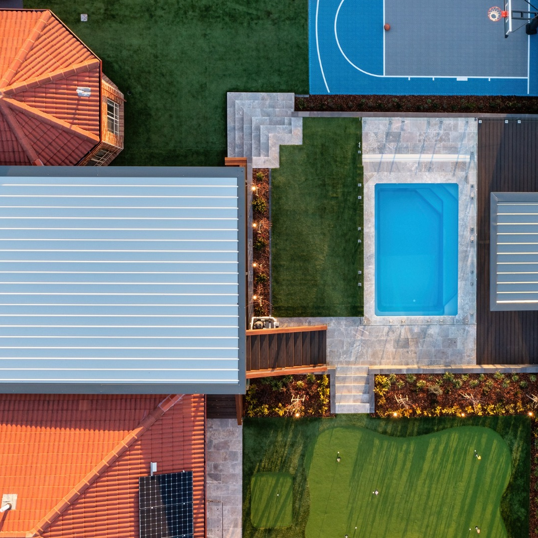 Aerial view of a residential backyard with a pool, sports court, putting green, and house with a terra-cotta roof.