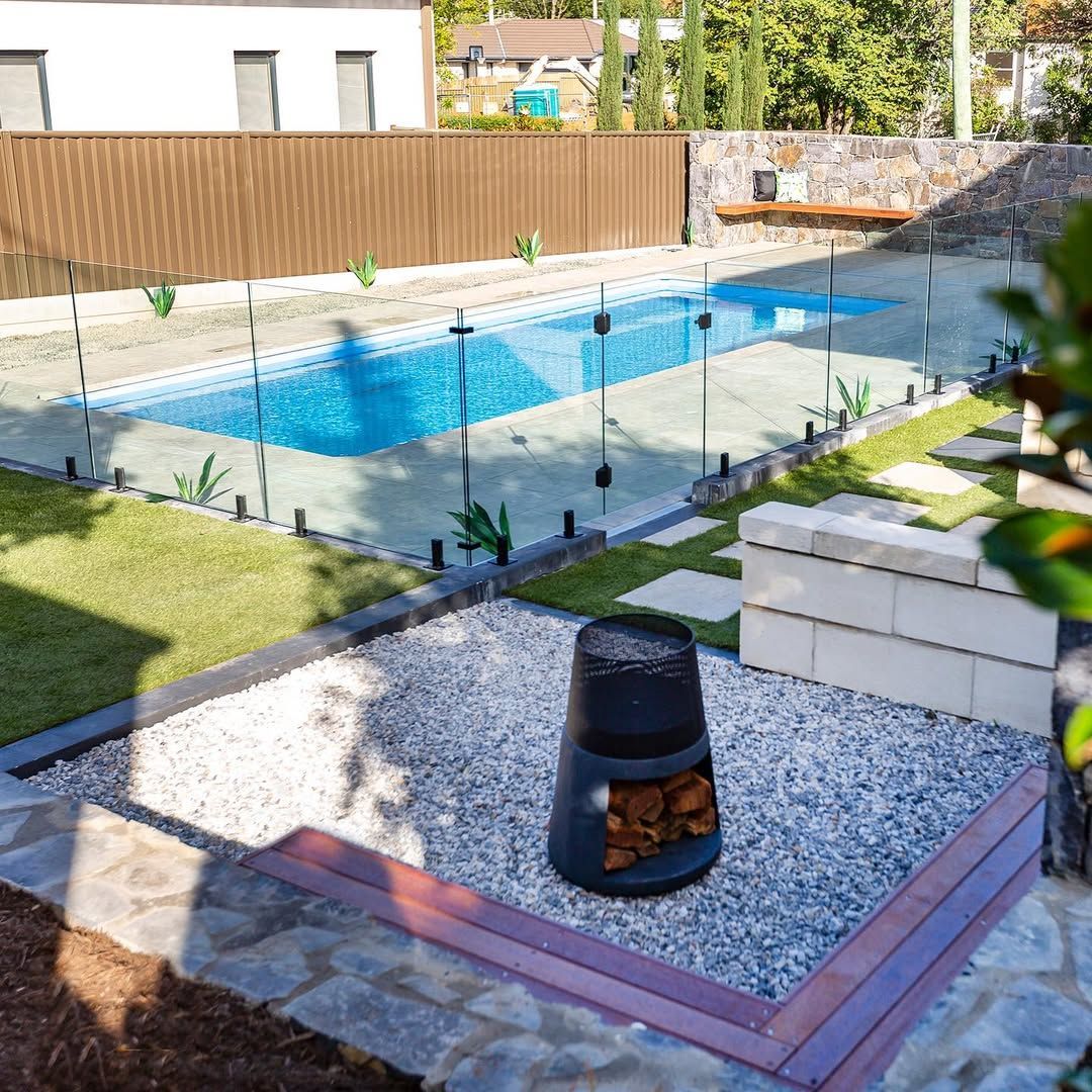 A backyard featuring a swimming pool, glass fencing, a gravel patio area with a fire pit, and a stone retaining wall.
