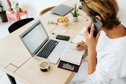 A person in a white shirt sits at a light wood table, talking on a smartphone while writing in a notebook near a laptop.