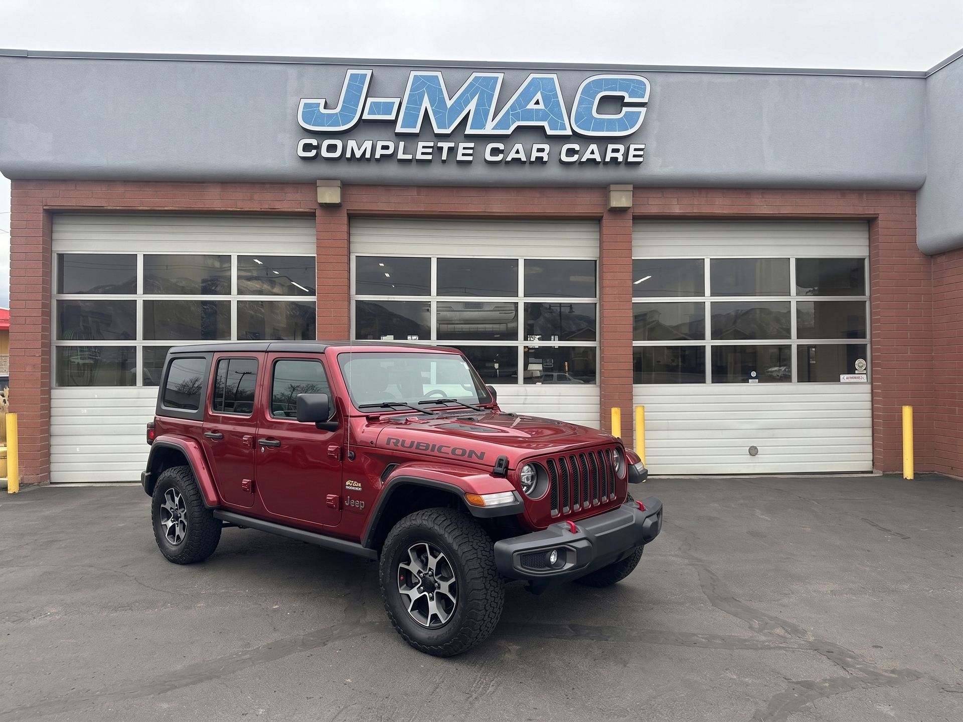Red Jeep Wrangler parked outside J-Mac Complete Car Care garage with closed bay doors
