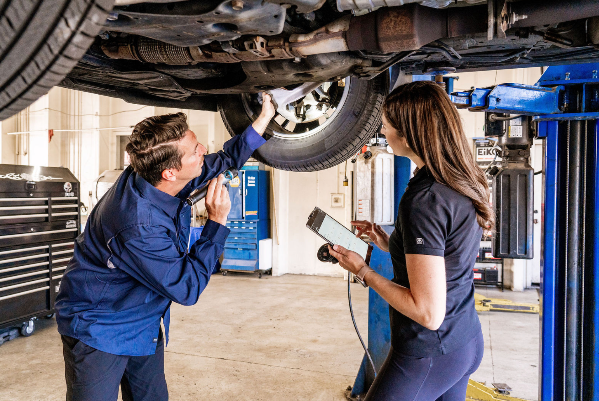 Mechanic inspecting a car on a lift while a technician with a clipboard observes in a garage