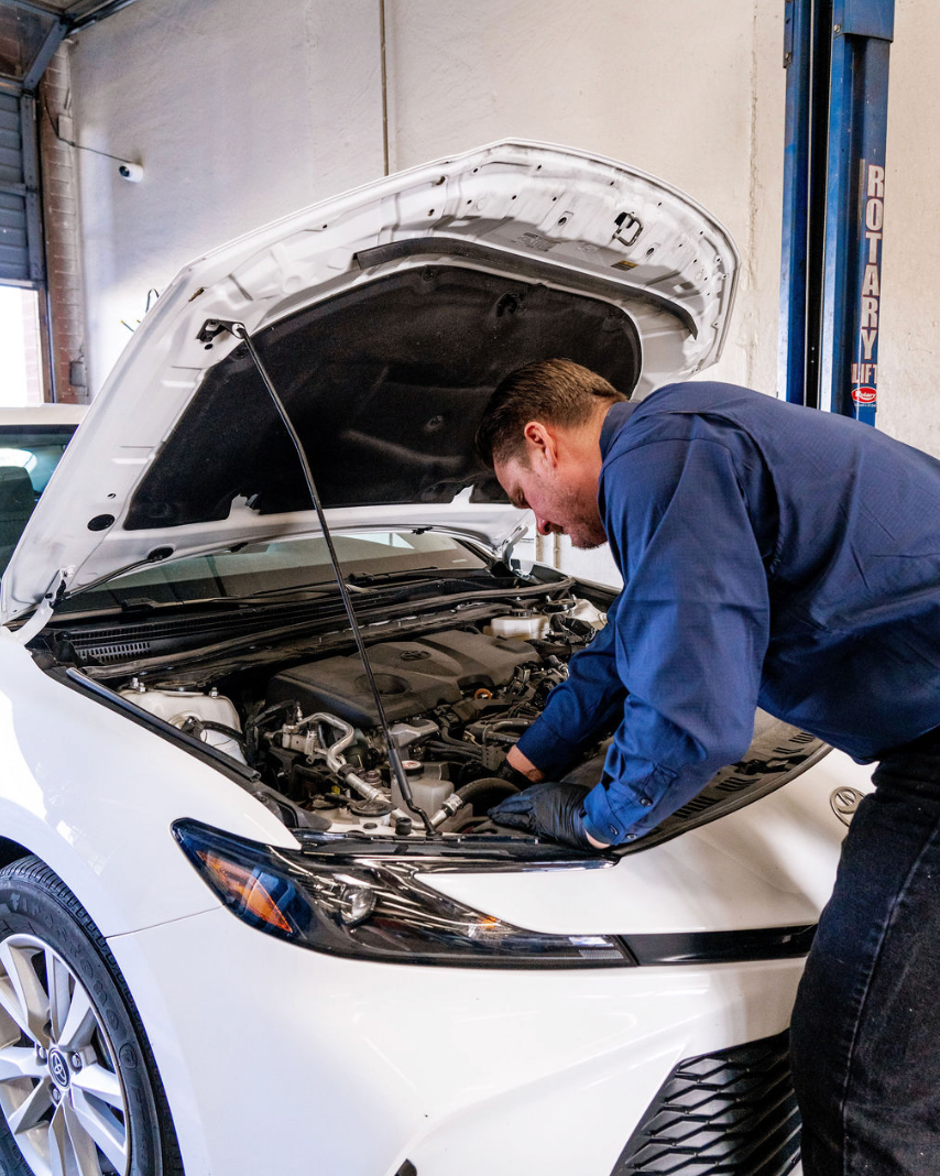 Mechanic in a blue shirt inspecting the engine of a white car with the hood open in a garage