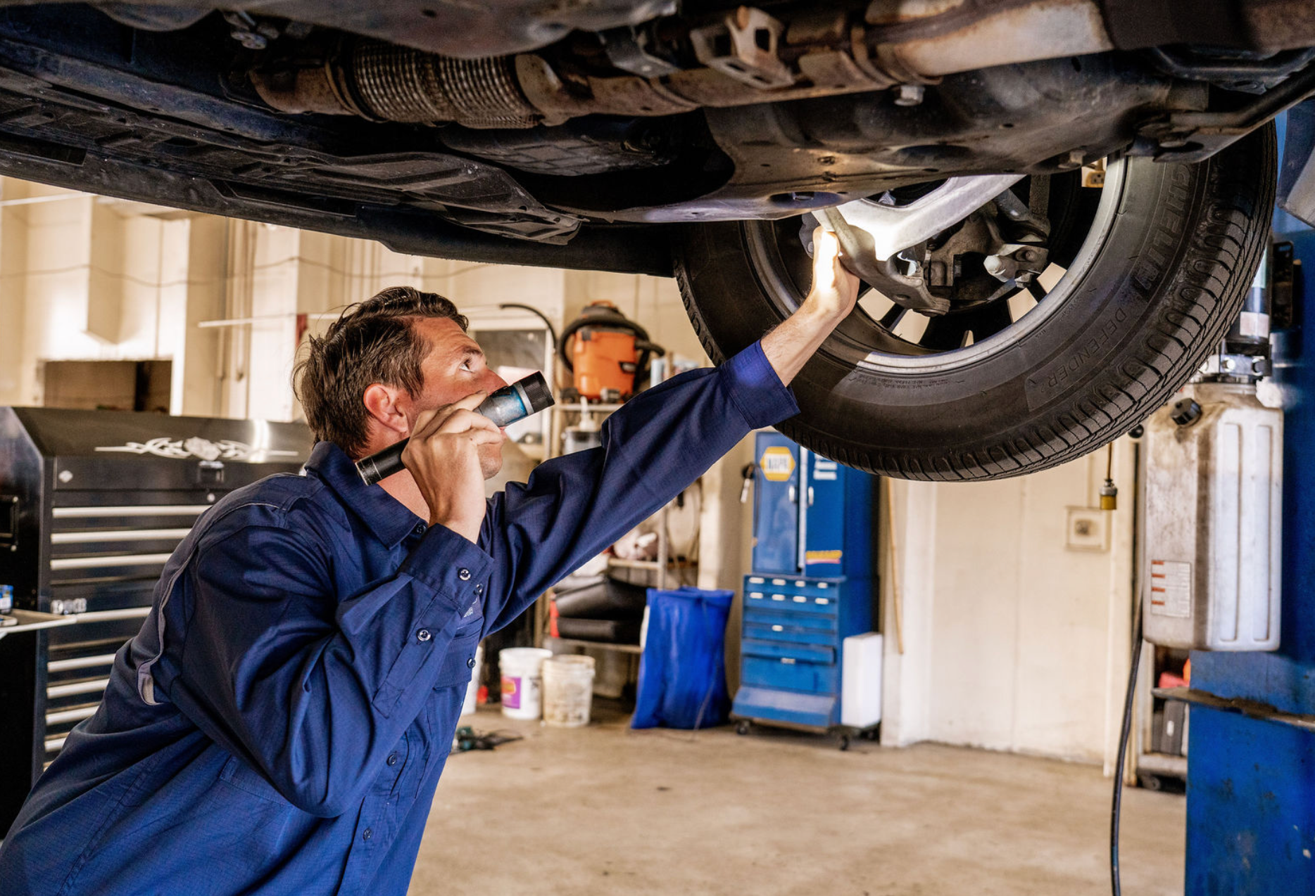 Mechanic inspecting a car’s undercarriage in a garage, wearing a blue uniform and gloves