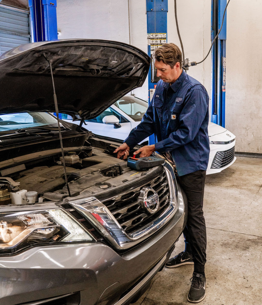 Mechanic inspecting a silver Nissan SUV with the hood open in an auto shop