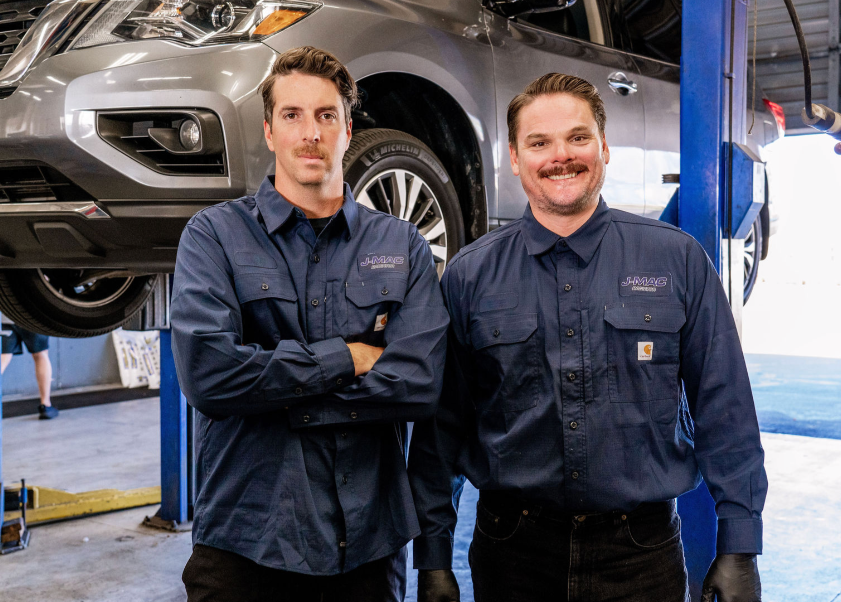 Two mechanics in navy uniforms stand in a car repair shop beside a raised SUV.