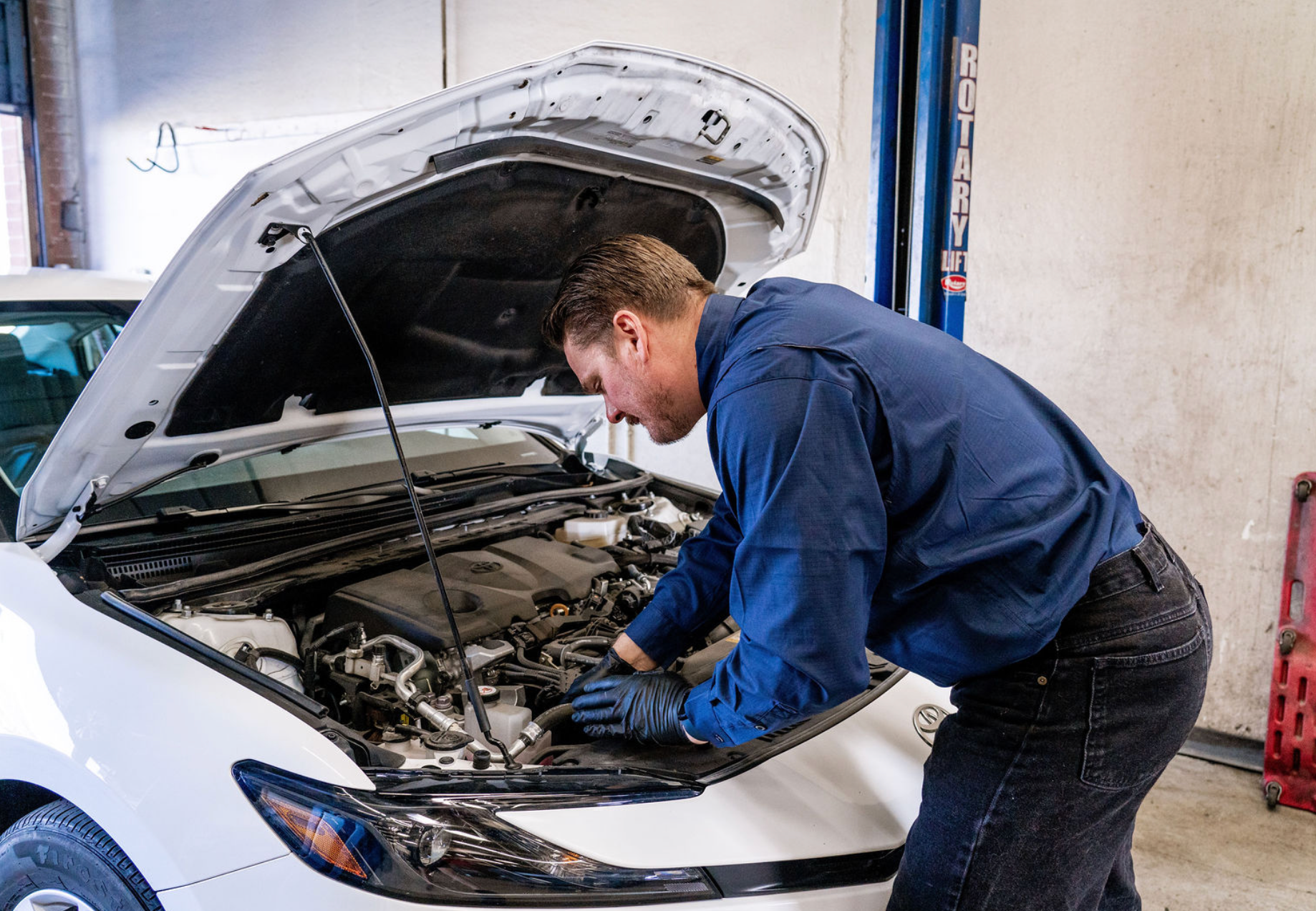Mechanic in blue coveralls inspecting a white car with its hood open in a garage