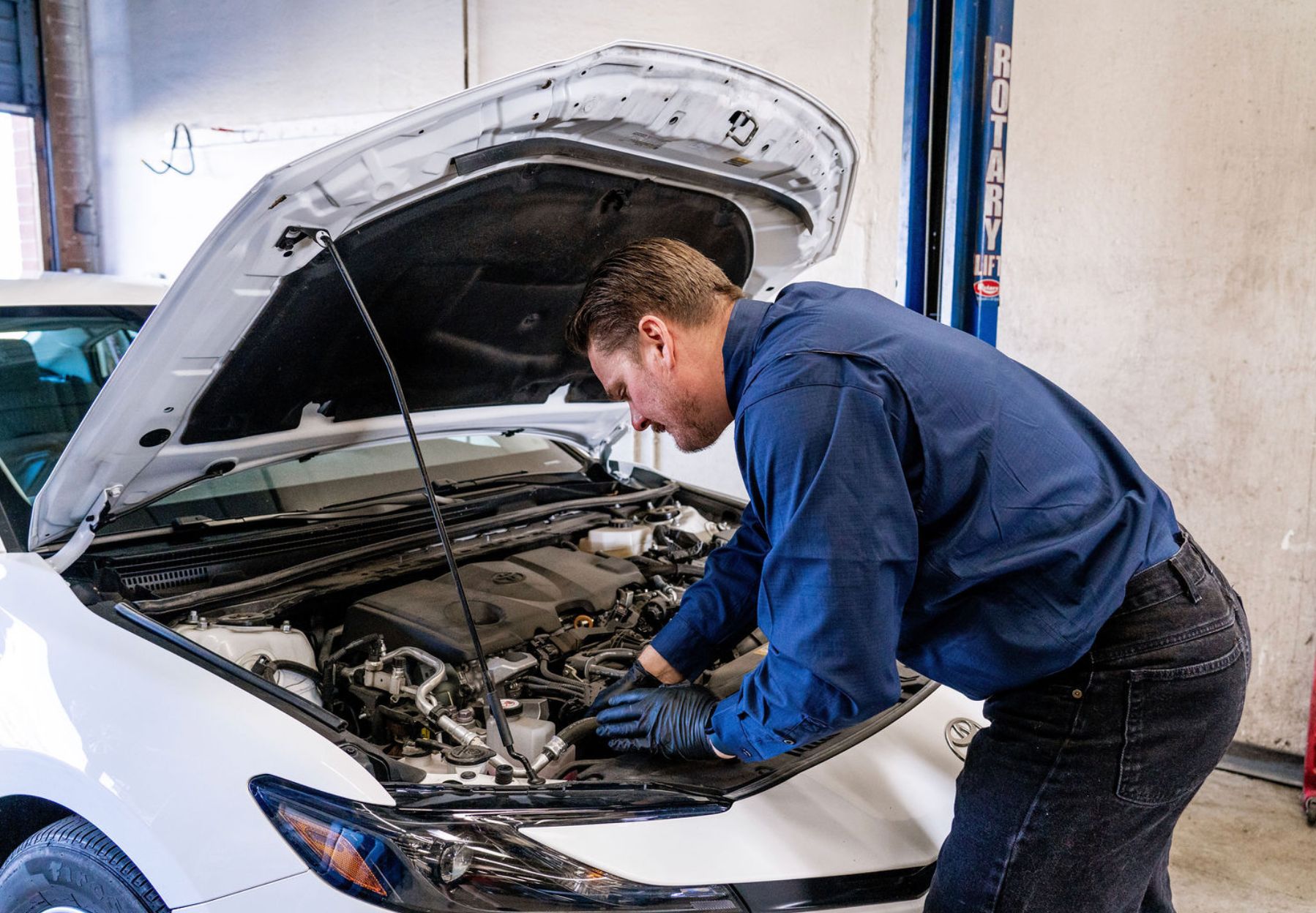 Mechanic in blue coveralls inspecting a white car engine with the hood open in a garage