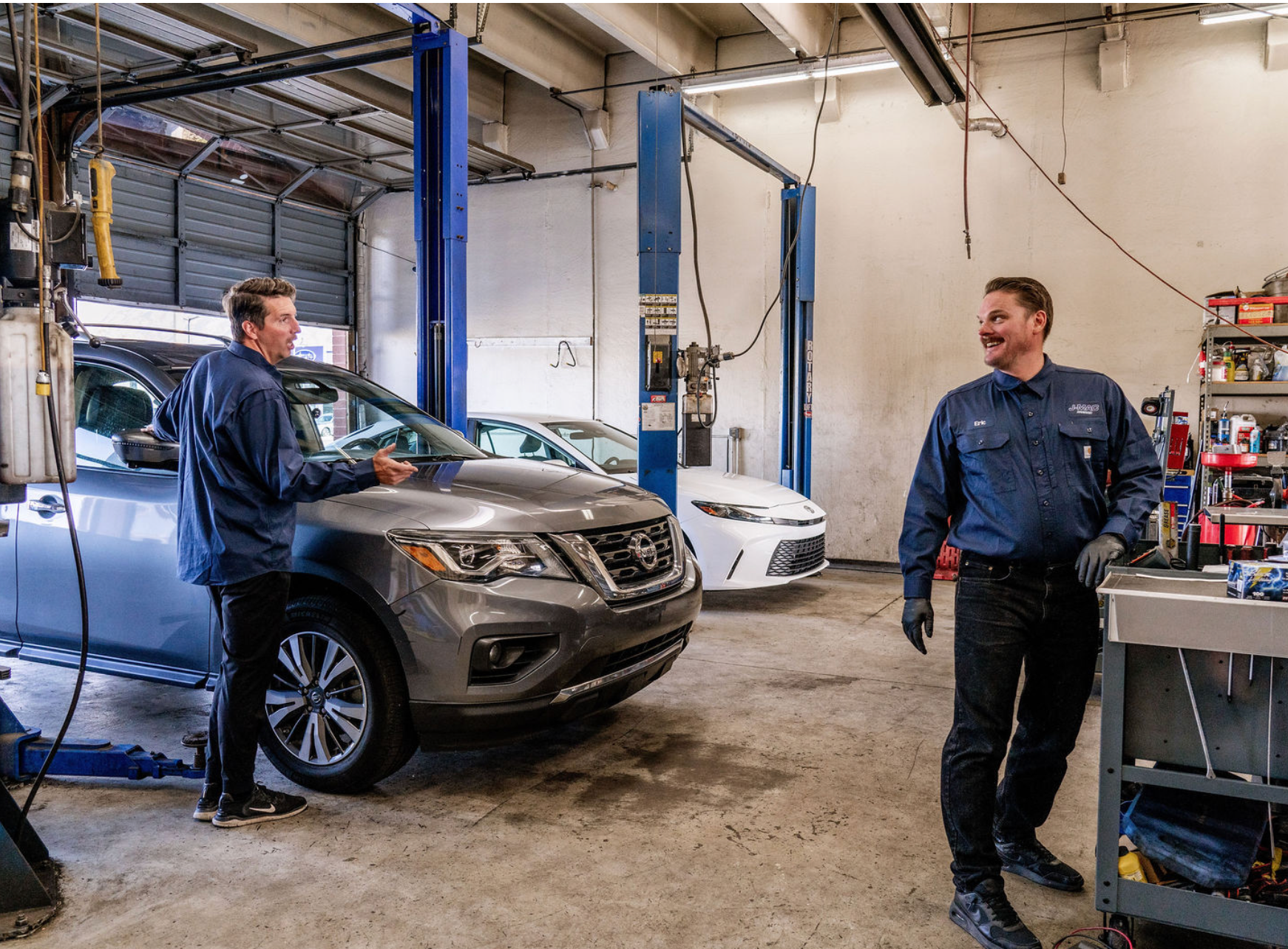 Two mechanics talking beside a silver SUV in an auto repair garage