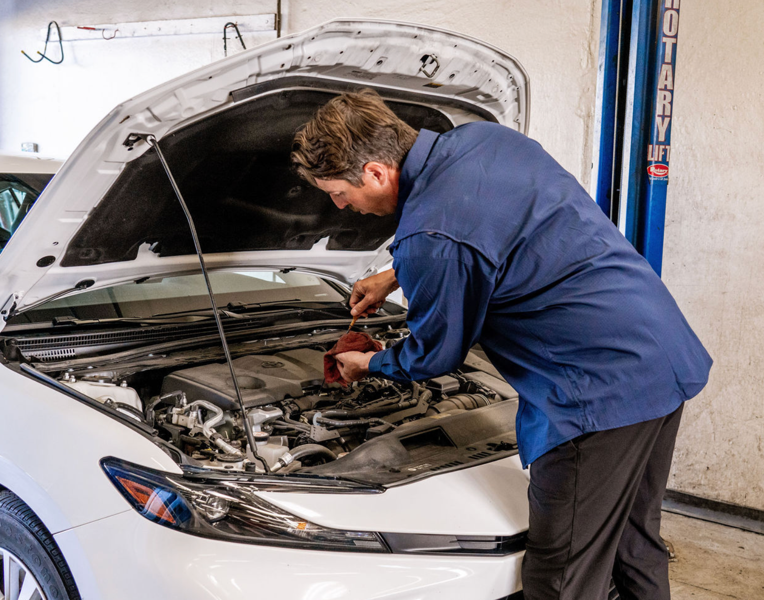 Mechanic in blue coveralls inspecting a white car’s engine with hood open in a repair shop