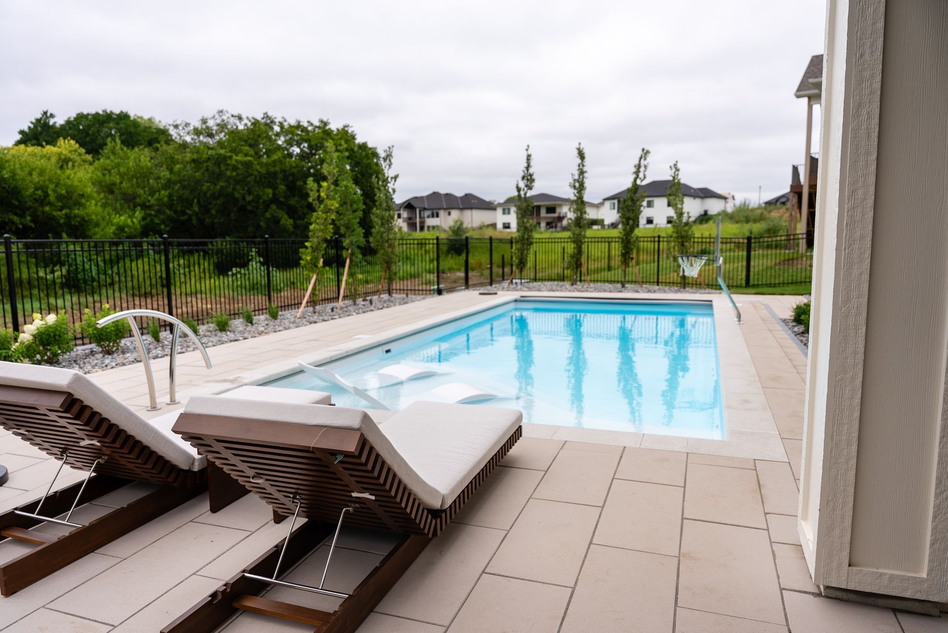 Rectangular pool with lounge chairs on a patio, overlooking a backyard with trees and houses.