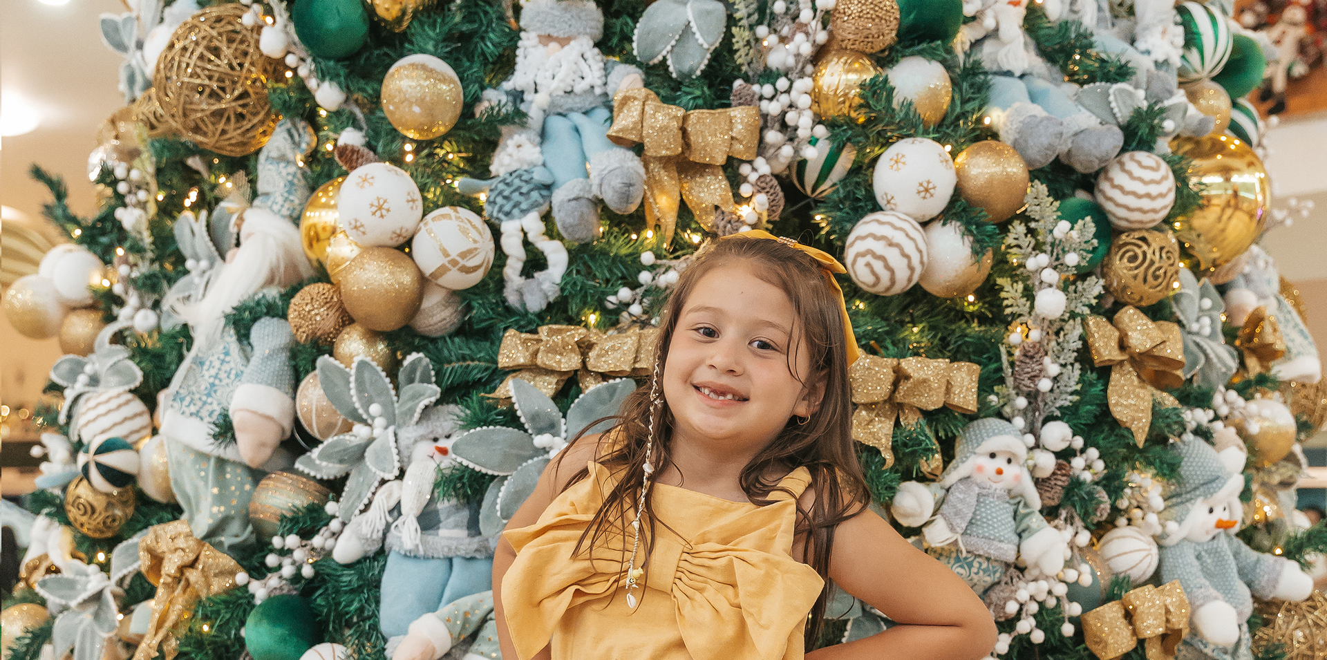 Menina sorridente de vestido amarelo posando em frente a um cenário festivo de Natal em branco, dourado e verde.