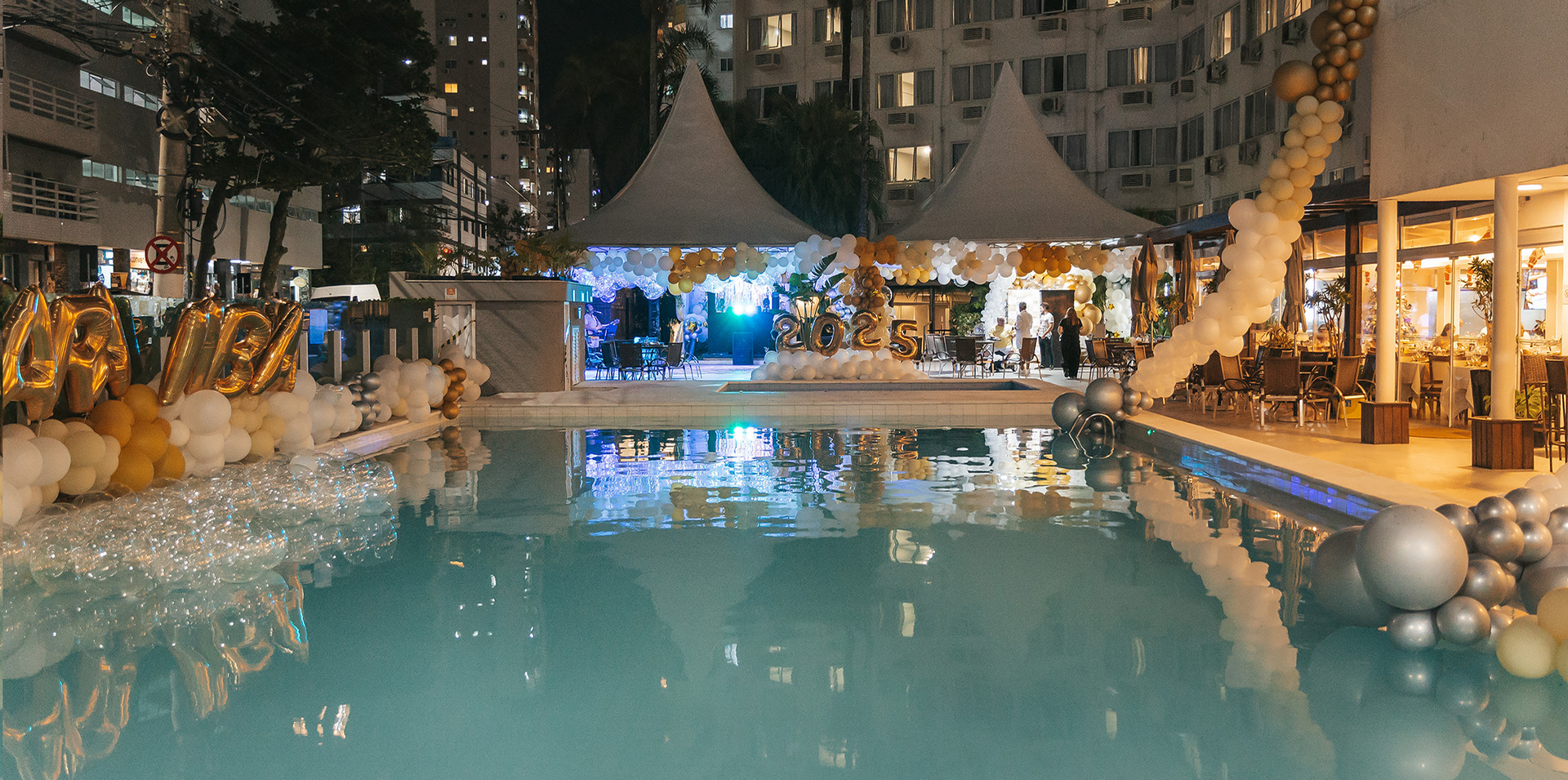 Resort à beira da piscina ao ar livre, coberto de neve, à noite, com luzes aconchegantes e um pavilhão central festivo refletido na água.