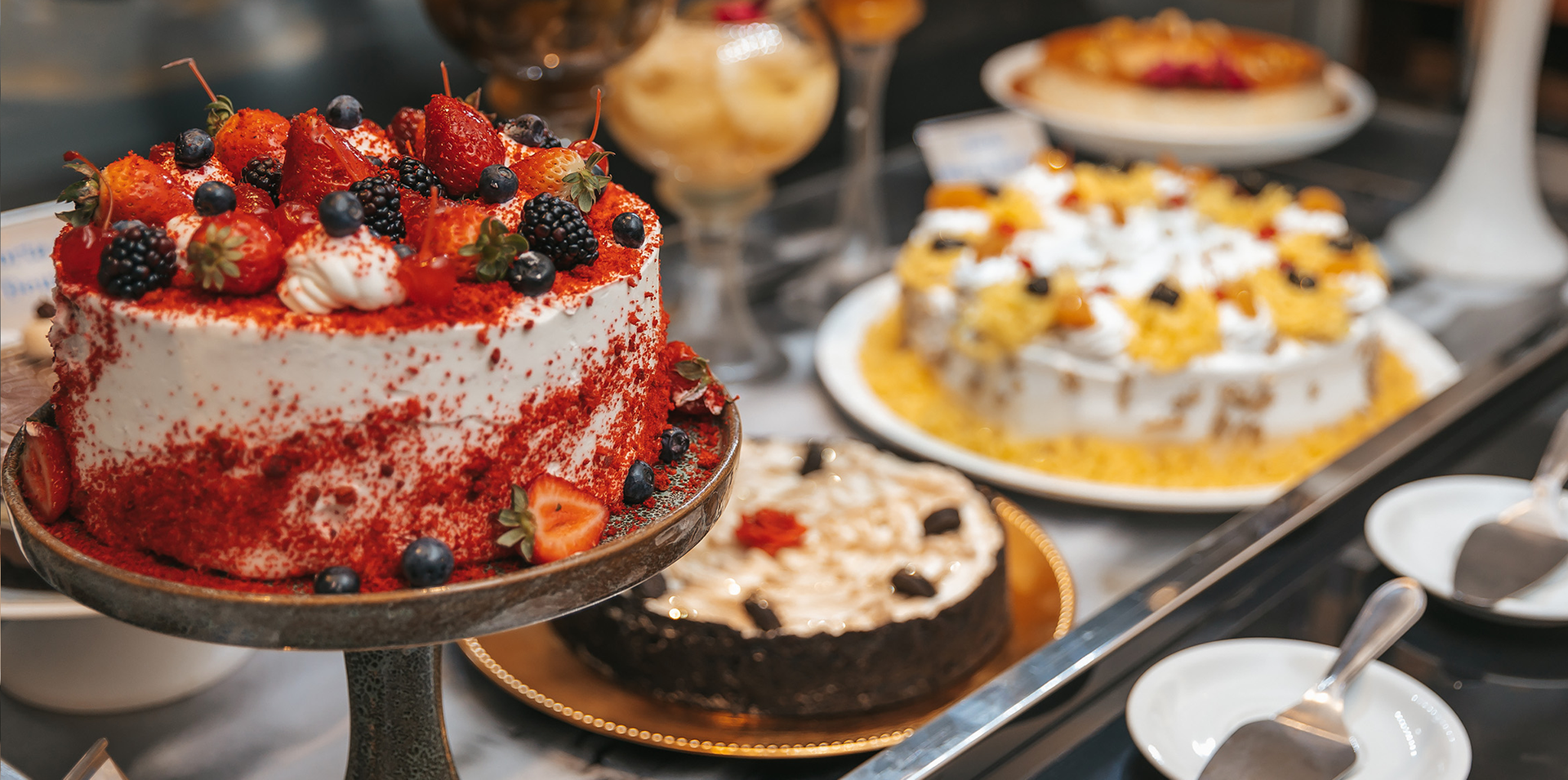 Diversos bolos decorados em suportes numa mesa de sobremesas, incluindo um bolo de frutos vermelhos e bolos com chantilly.