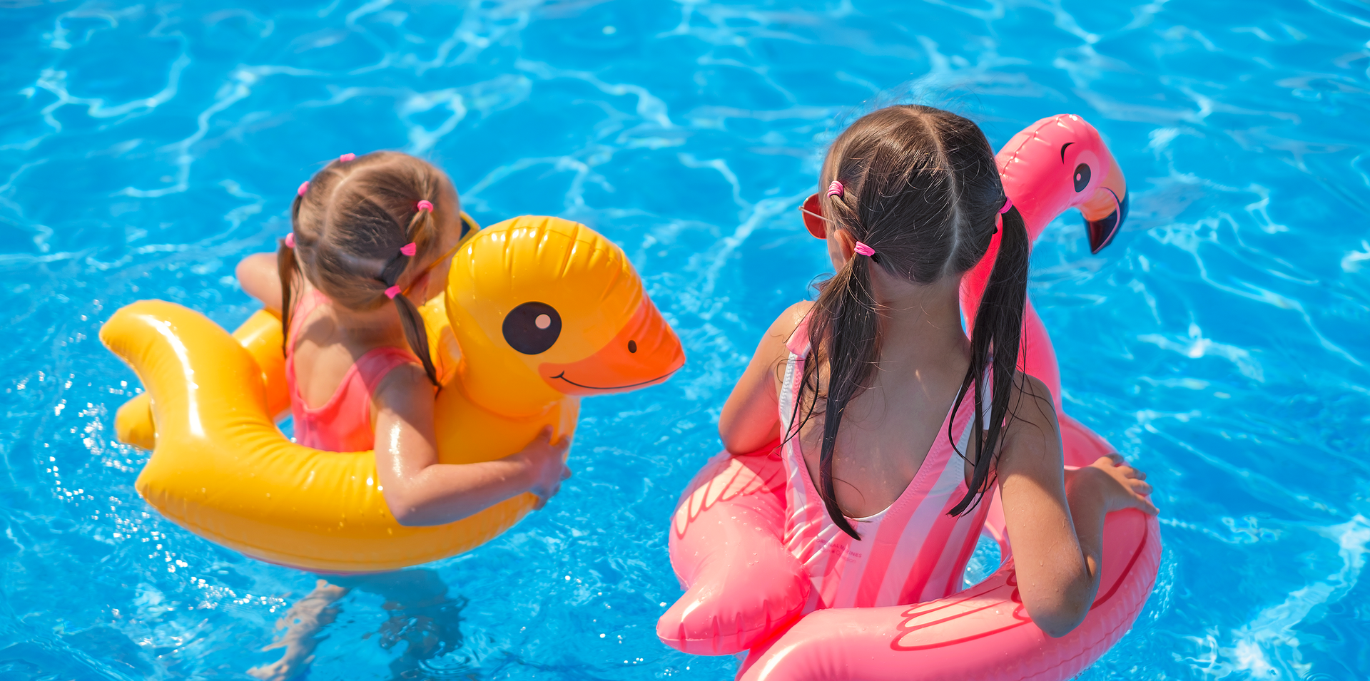 Duas crianças flutuando em boias infláveis coloridas em formato de cisne em uma piscina azul brilhante.