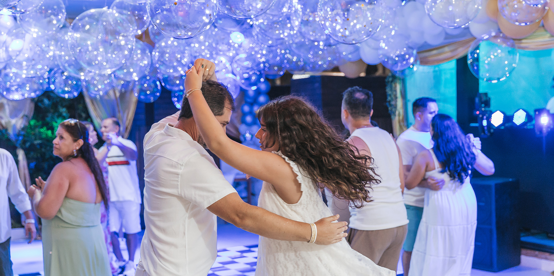 Convidados dançando sob uma decoração de balões azuis e brancos em uma festa animada em ambiente fechado.