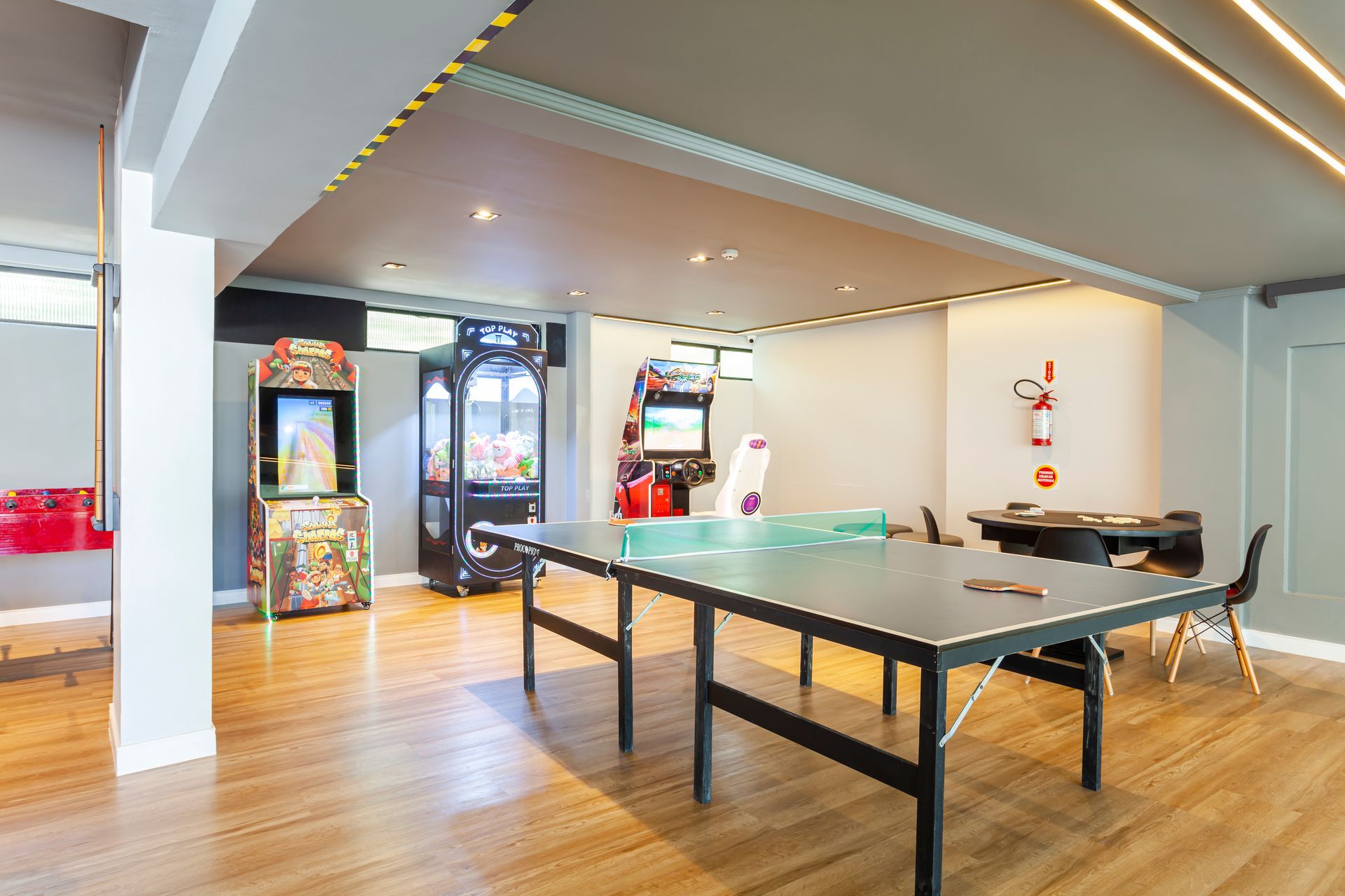 Sala de jogos moderna com mesa de pingue-pongue, máquinas de fliperama e banquetas de bar em piso de madeira.