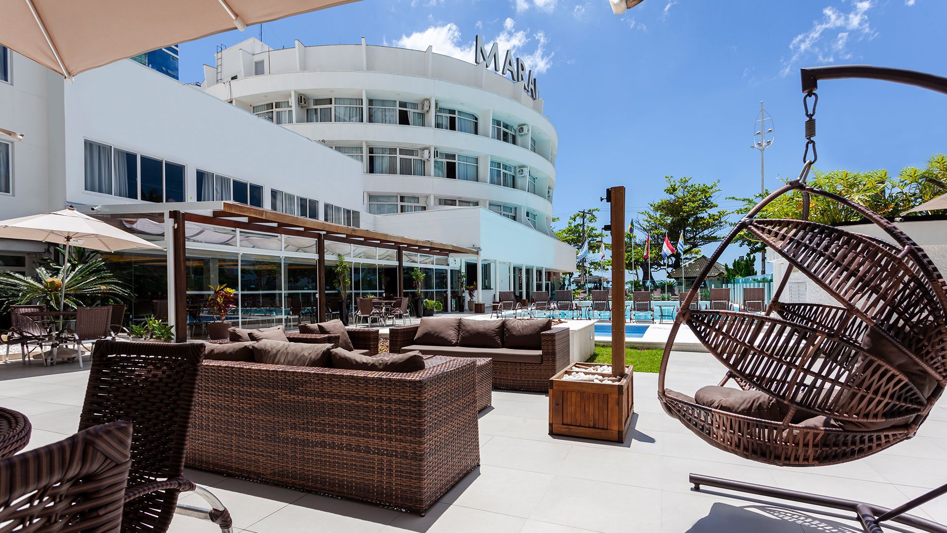 Terraço no terraço do resort com espreguiçadeiras de vime, piscina e prédio curvo branco do hotel sob um céu azul.