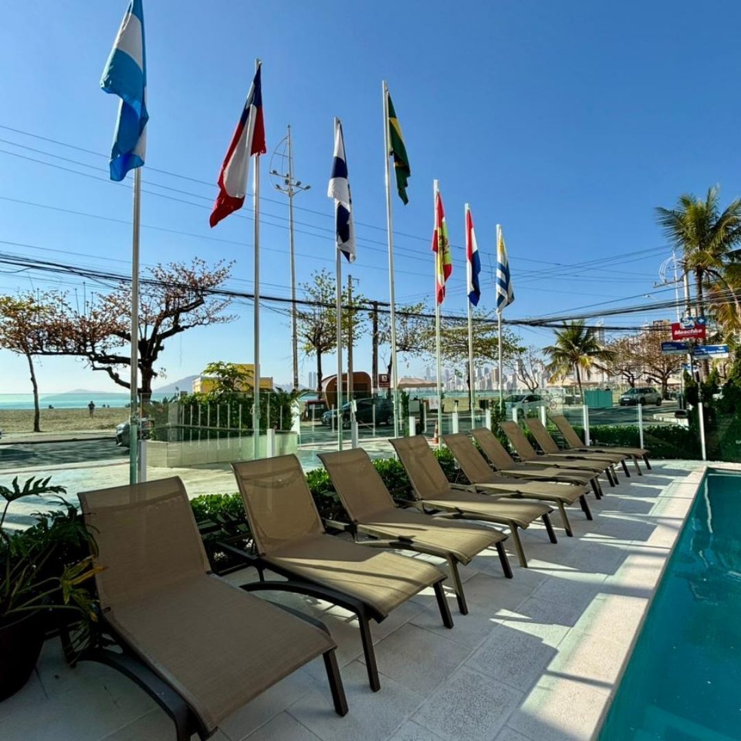 Espreguiçadeiras à beira de uma piscina ensolarada com bandeiras e o oceano ao fundo.