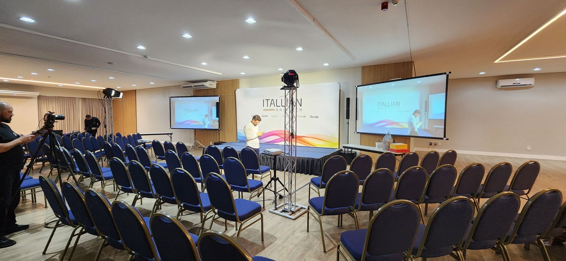 Sala de conferências com fileiras de cadeiras azuis, um pódio e telas de projeção na frente.