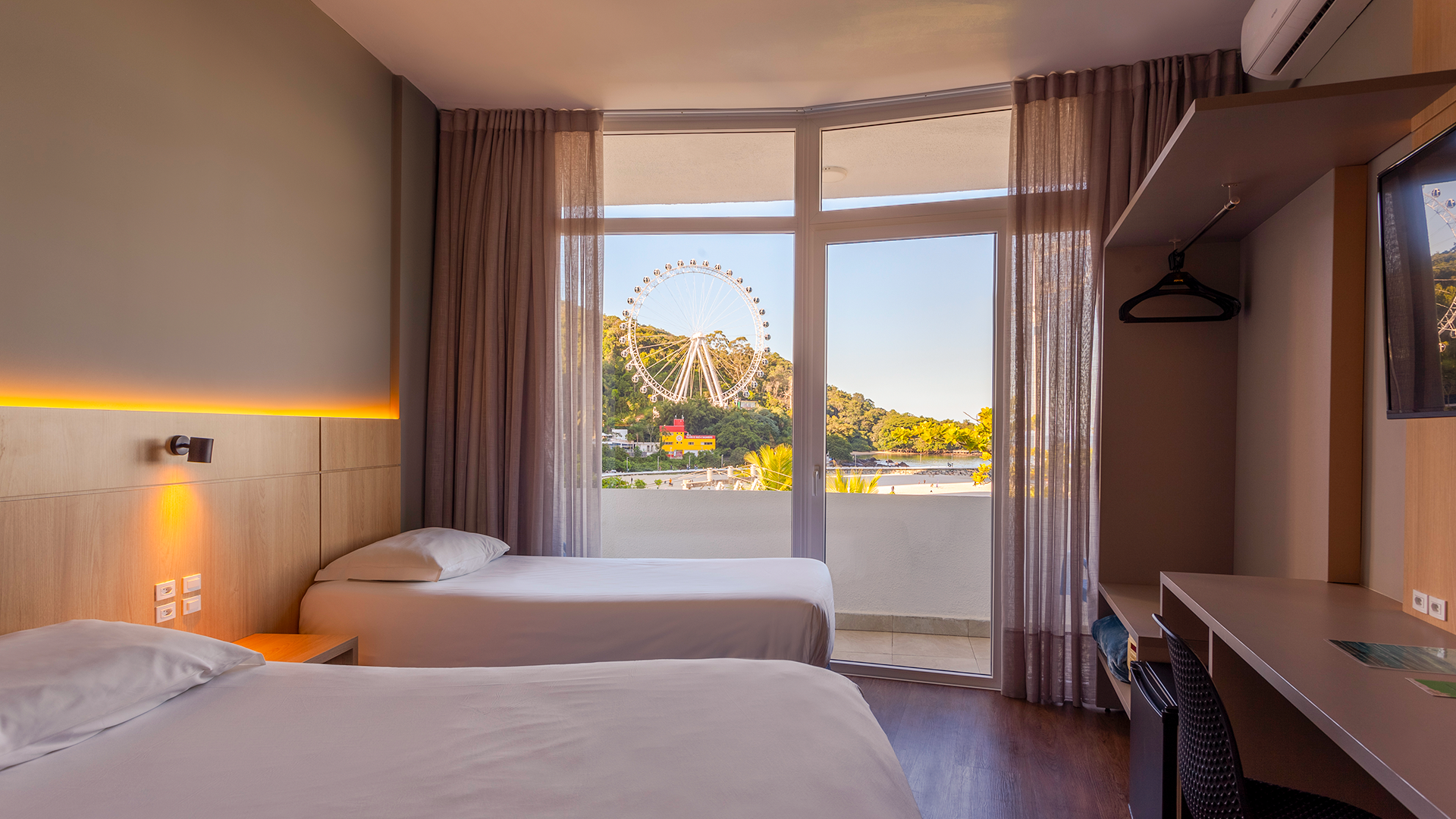 Quarto de hotel luminoso com duas camas, uma escrivaninha e amplas portas de varanda com vista para a roda-gigante à beira-mar.