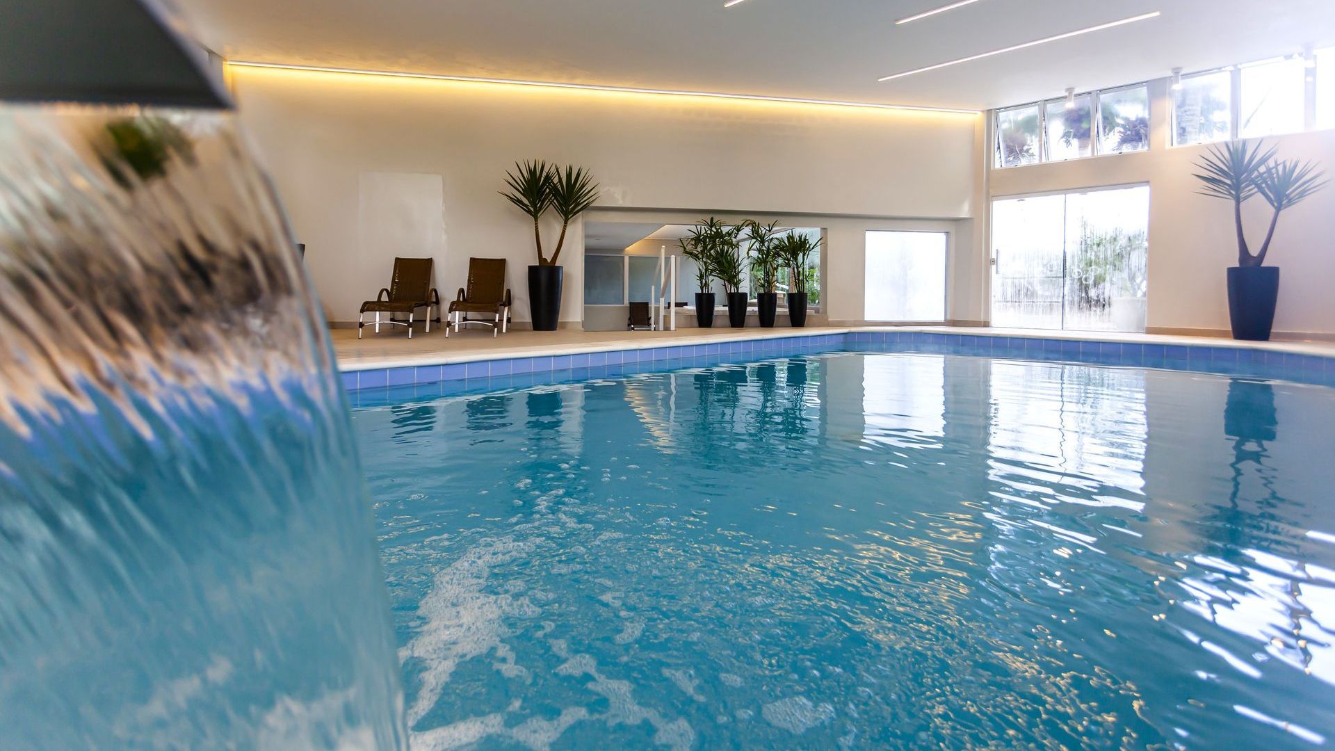 Piscina coberta com água azul, espreguiçadeiras e grandes janelas em um ambiente luminoso que lembra um spa.