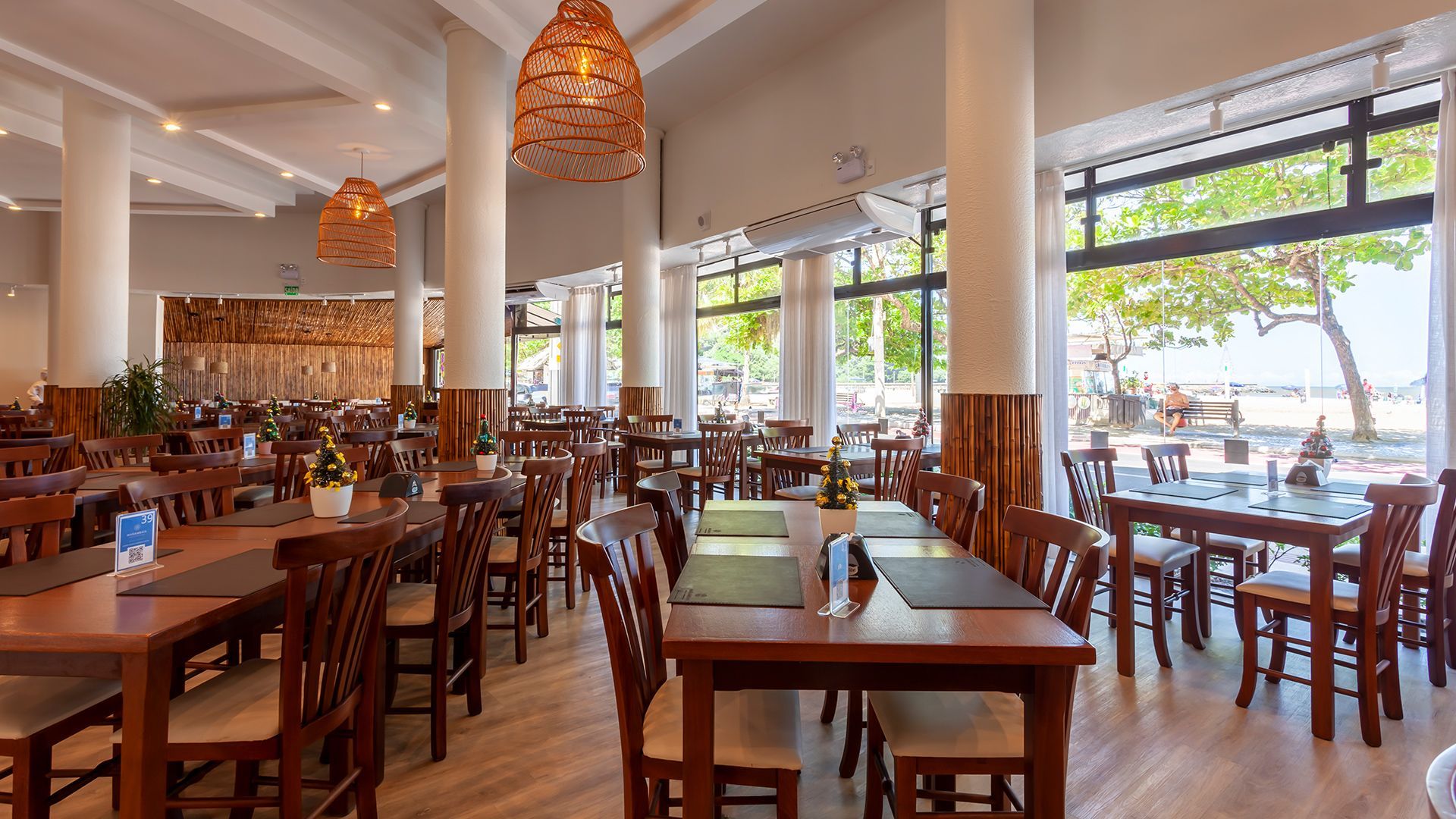 Restaurante ao ar livre com mesas e cadeiras de madeira, luminárias pendentes de vime e janelas com vista para o mar.