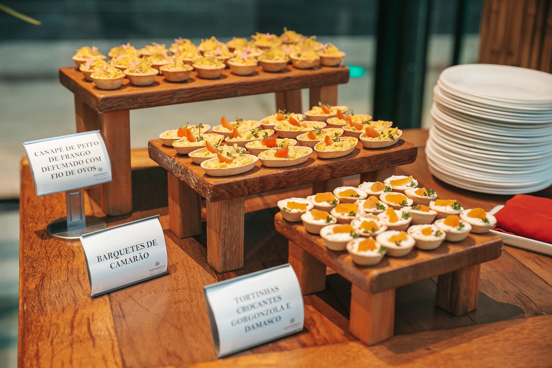 Buffet de sobremesas com mini doces em suportes de madeira ao lado de pratos brancos empilhados e cartões de lugar.
