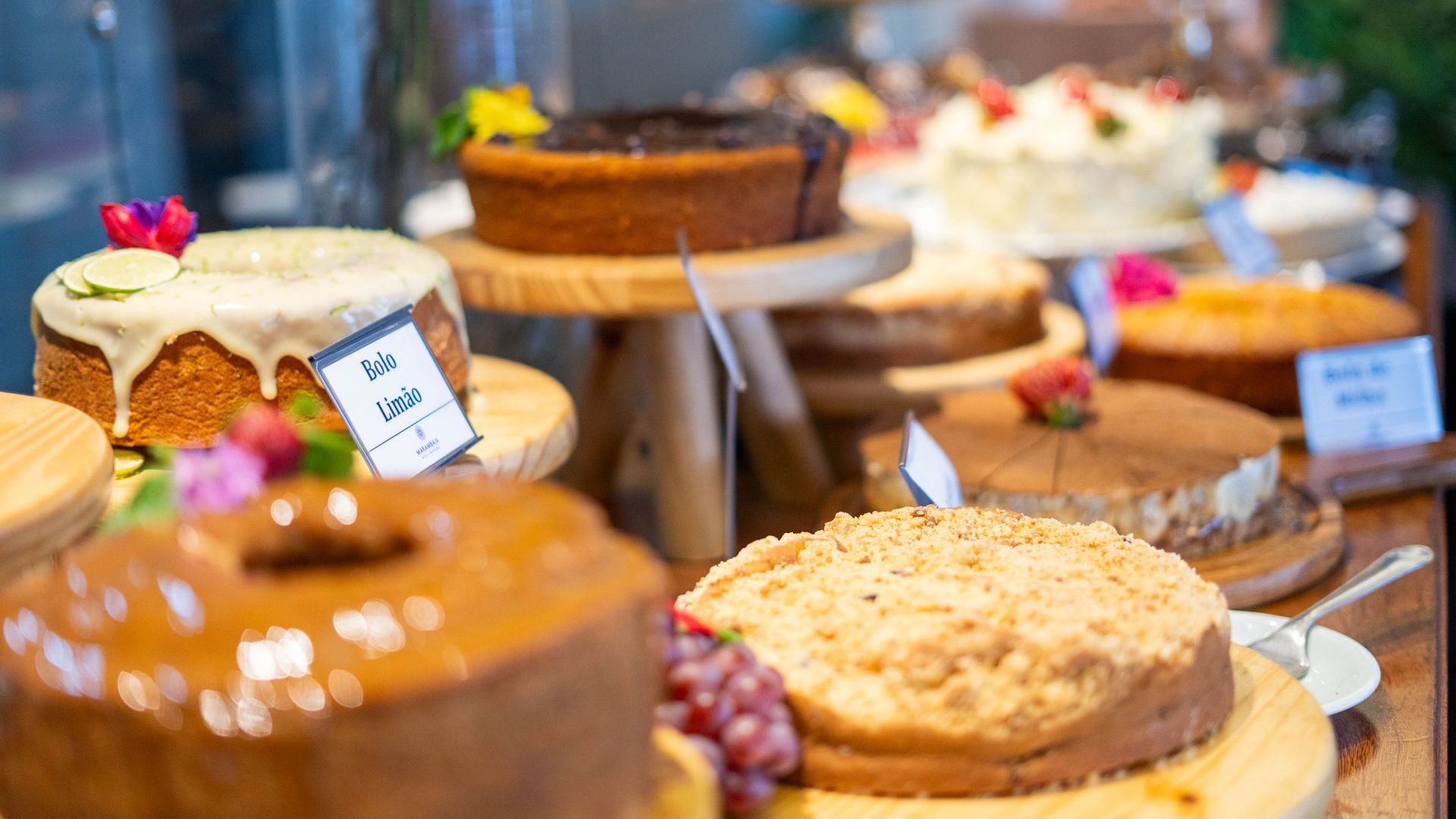 Bolos e doces variados expostos em uma mesa de bufê com pequenas etiquetas.