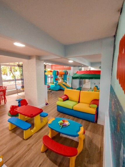 Área de recreação interna colorida com mesas e cadeiras infantis, além de um sofá amarelo ao lado de uma piscina de bolinhas.