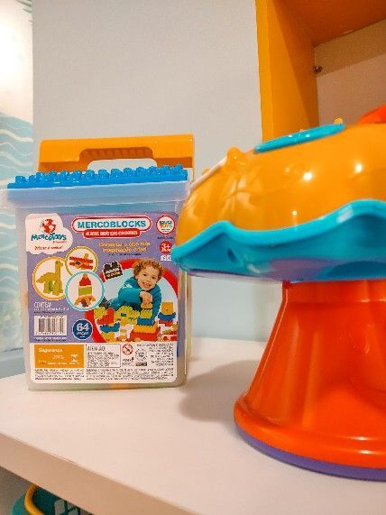 Brinquedo de plástico colorido sobre uma mesa ao lado de um conjunto de brinquedos infantil embalado em uma caixa, em uma sala com paredes laranja.