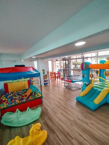 Área de recreação interna colorida com piscina de bolinhas, escorregadores e brinquedos em um ambiente iluminado com piso de madeira.