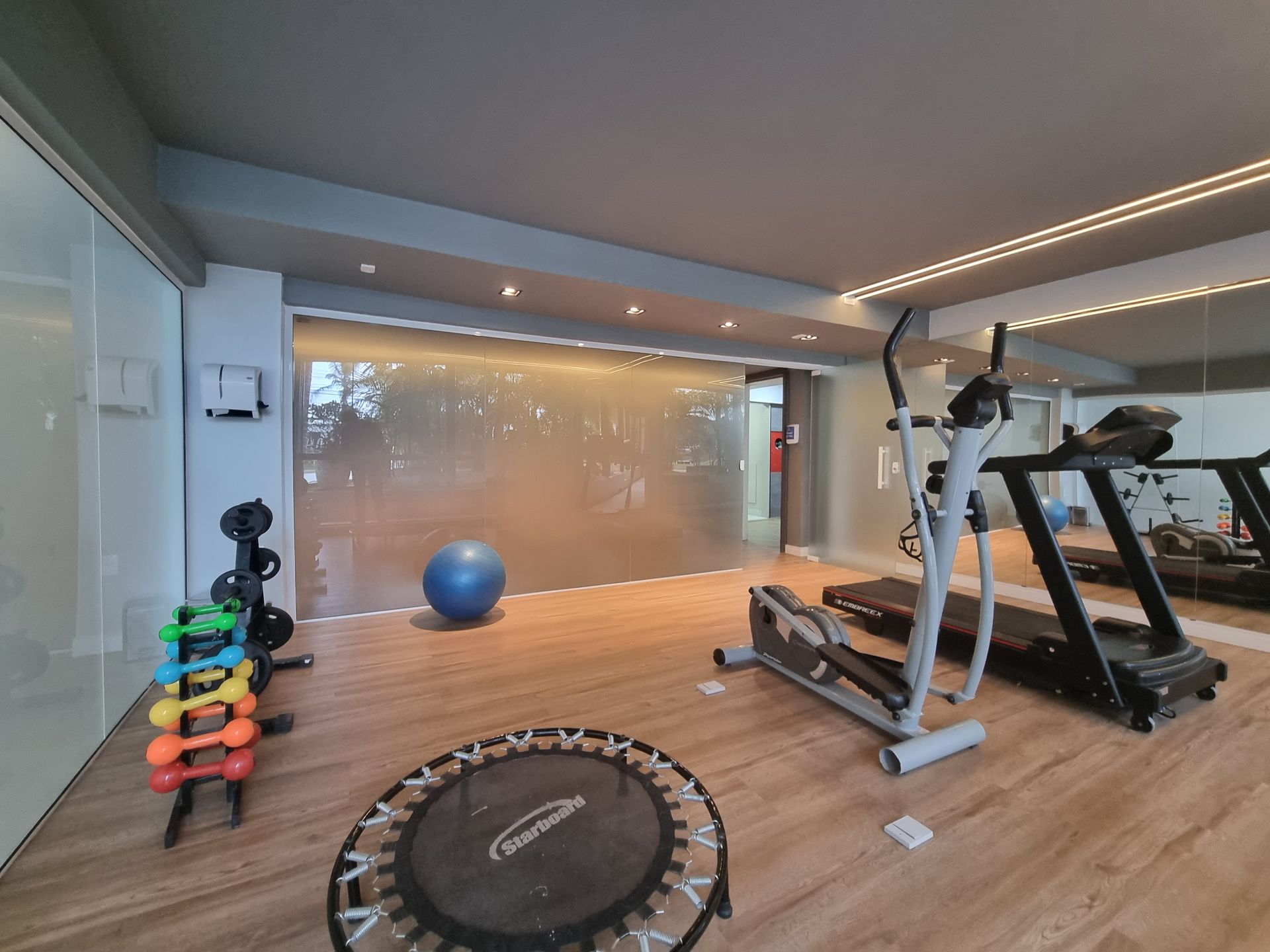 Academia moderna com esteira, bicicleta ergométrica, pesos, bola de estabilidade e cama elástica em piso de madeira.