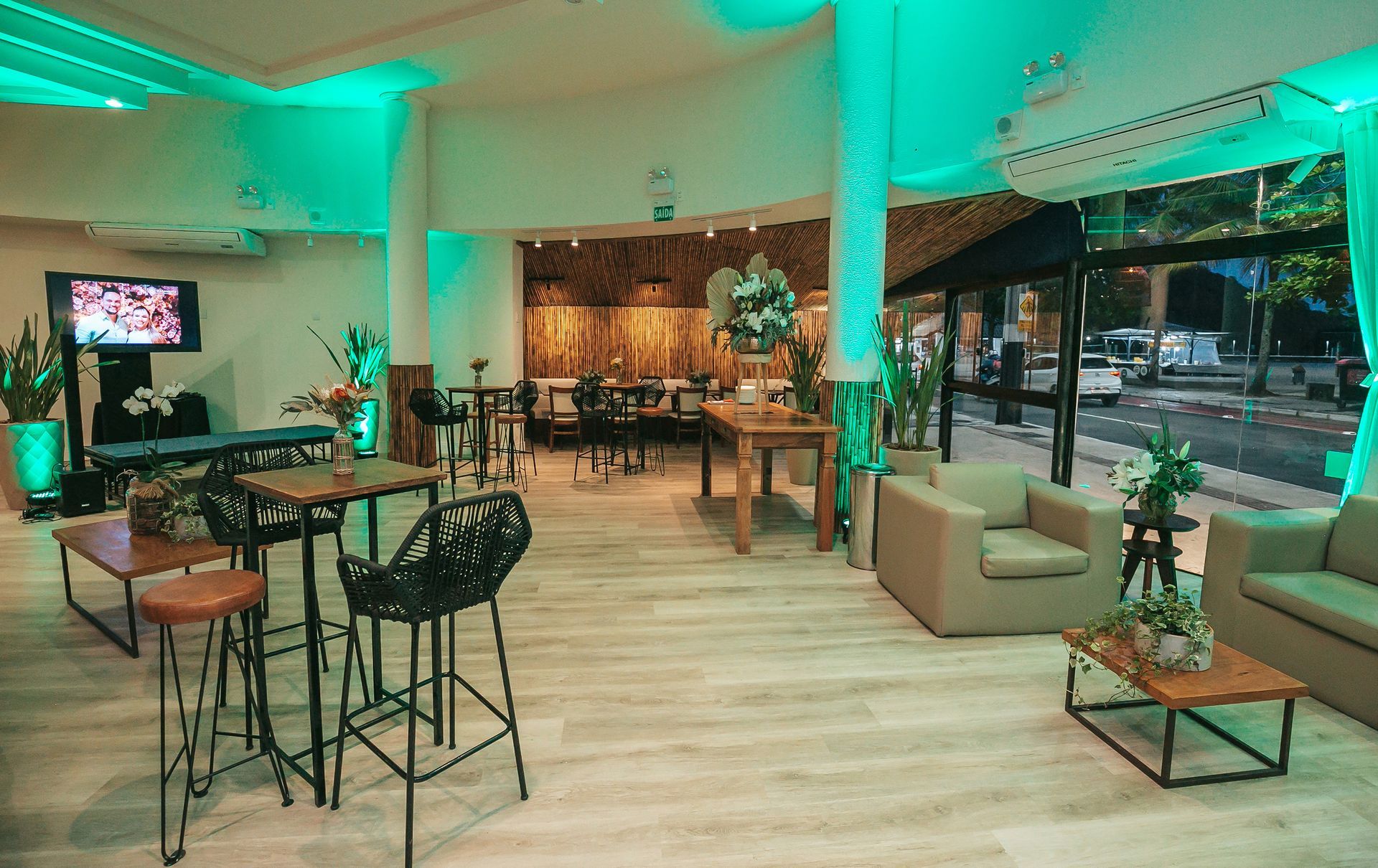 Sala de estar moderna com mesas altas, cadeiras e sofás sob iluminação verde, com vista para a rua.
