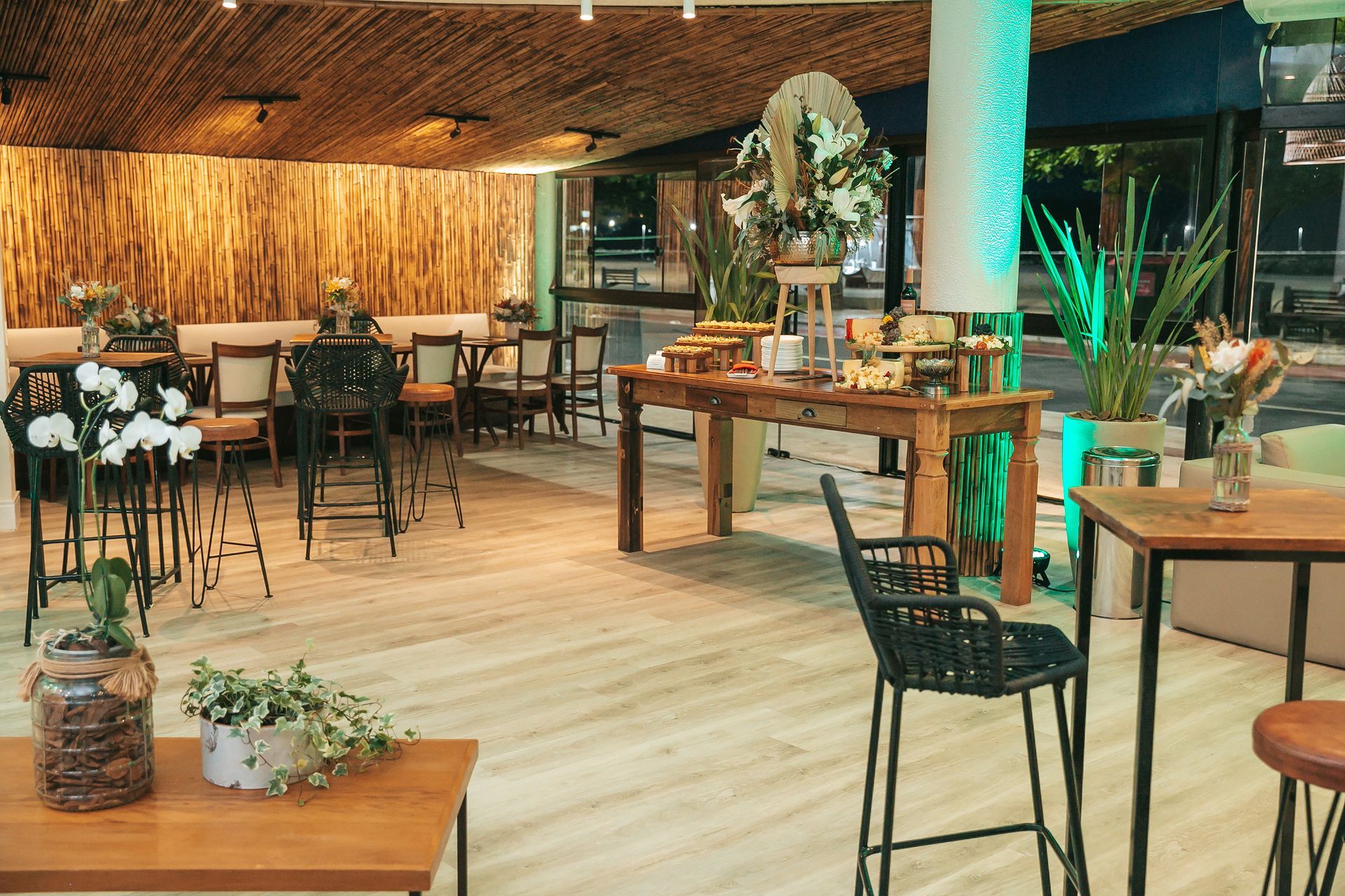 Interior acolhedor de restaurante tropical com cadeiras de vime, mesas de madeira e plantas em vasos.