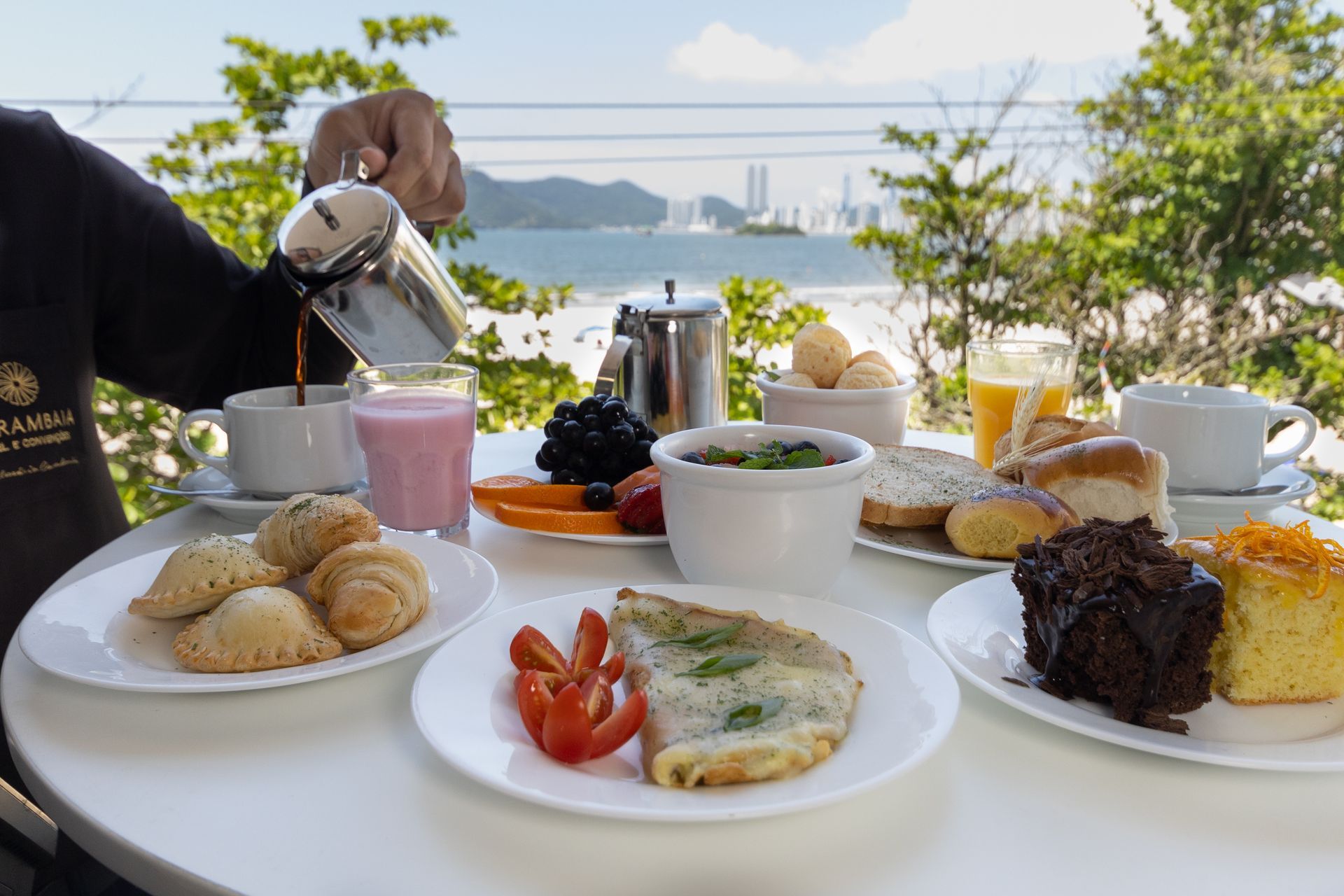 Mesa de café da manhã ao ar livre com café, pão, frutas e pratos servidos, com vista para a água.