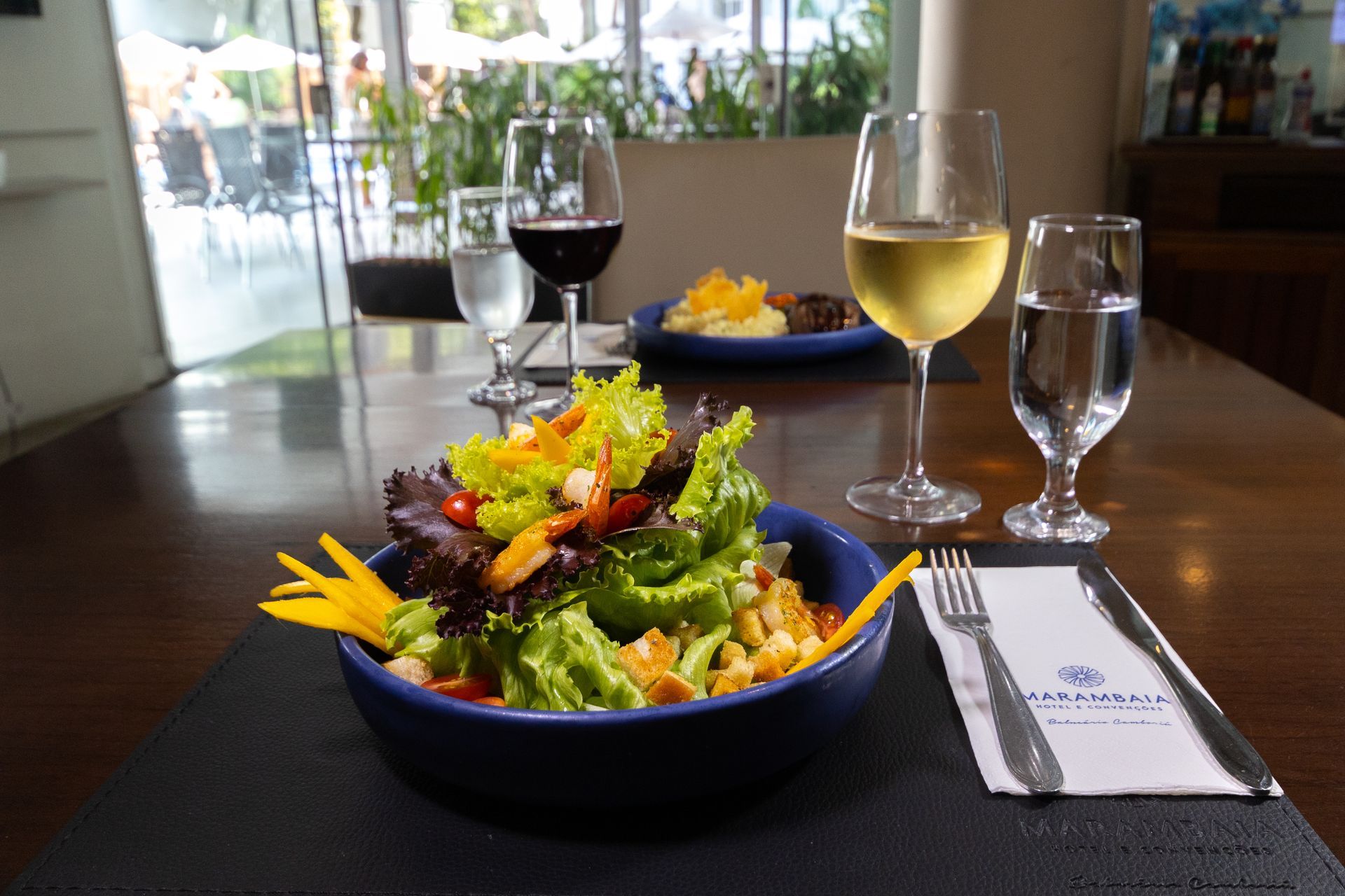 Mesa de restaurante com salada, taças de vinho e um copo de água junto a uma janela ensolarada.