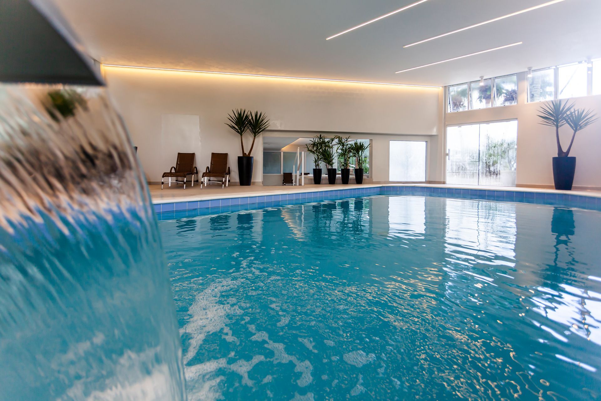 Piscina interior com água turquesa, espreguiçadeiras e grandes janelas num espaço luminoso com ambiente de spa.