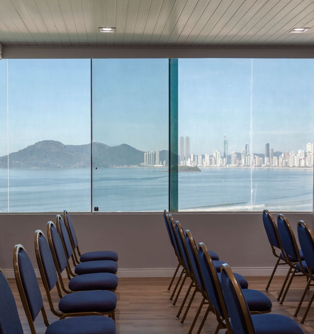 Fileiras de cadeiras azuis voltadas para grandes janelas com vista para a baía e o horizonte distante da cidade ao fundo.