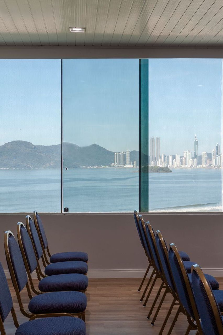 Sala de conferências vazia com fileiras de cadeiras voltadas para uma grande janela com vista para o mar e para o horizonte distante da cidade.