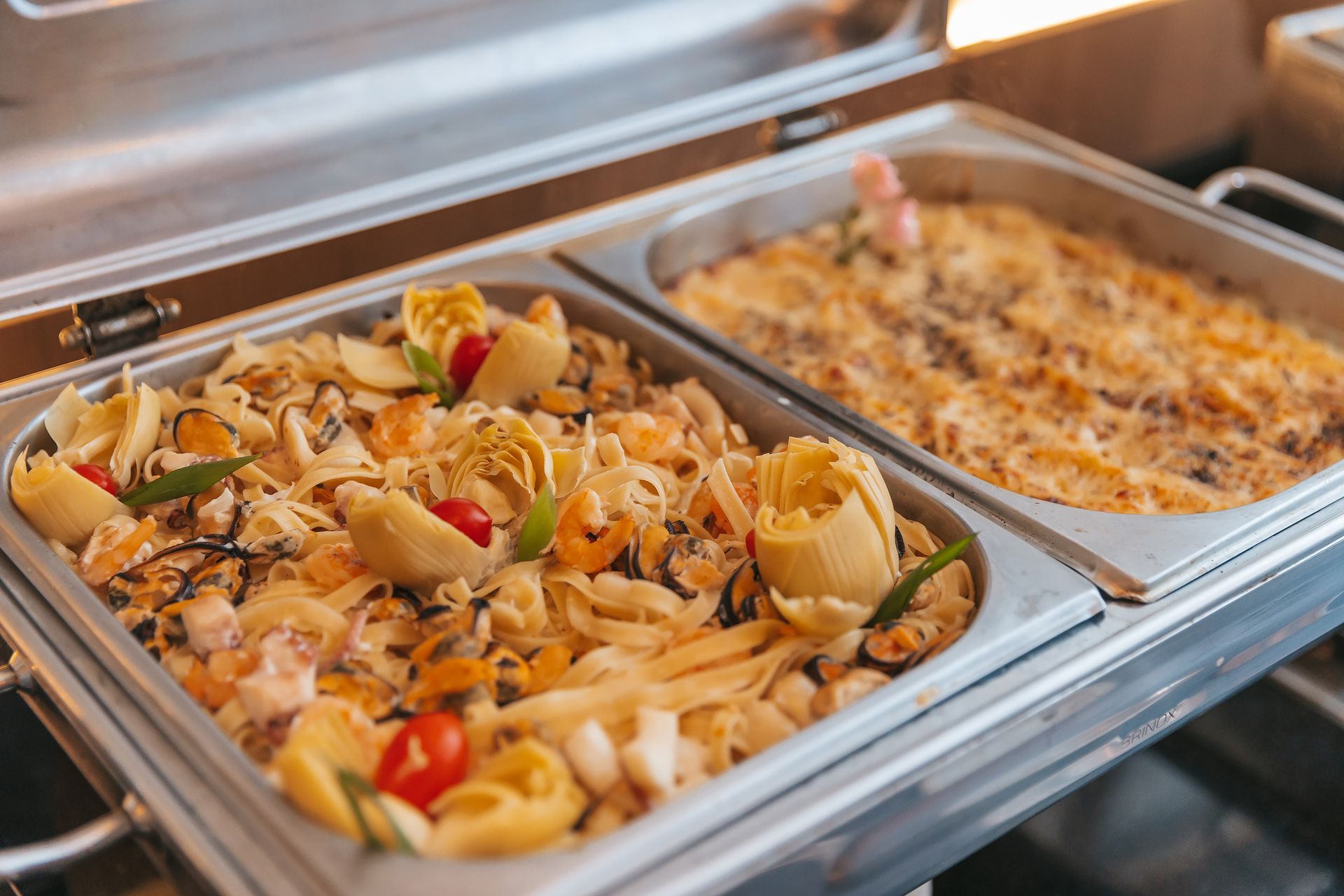 Bandeja com diversos pratos de massa em um balcão de buffet, incluindo massa com vegetais e massa assada.