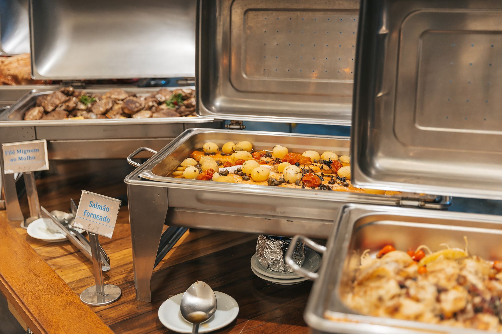 Mesa de buffet com bandejas cobertas contendo alimentos variados e utensílios para servir.