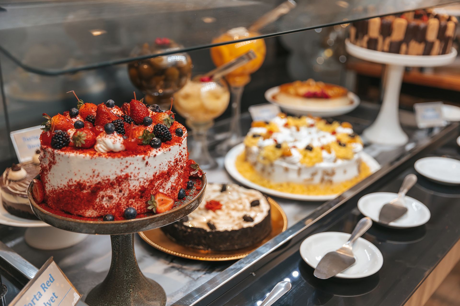 Vitrine de confeitaria com diversos bolos decorados em suportes e bandejas atrás de um vidro.