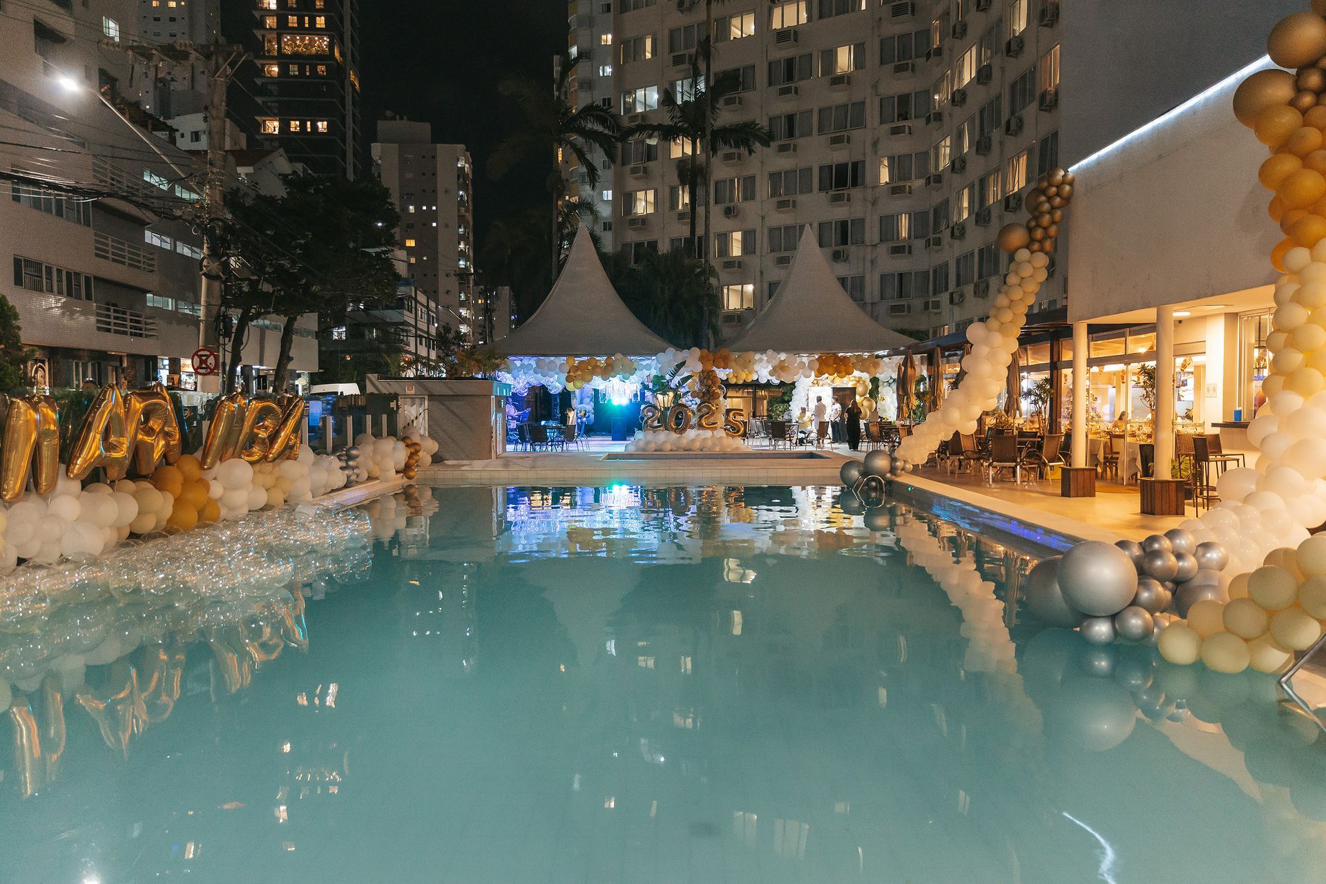 Piscina ao ar livre à noite, entre prédios altos, iluminada com água azul e luzes de corda em tons quentes.