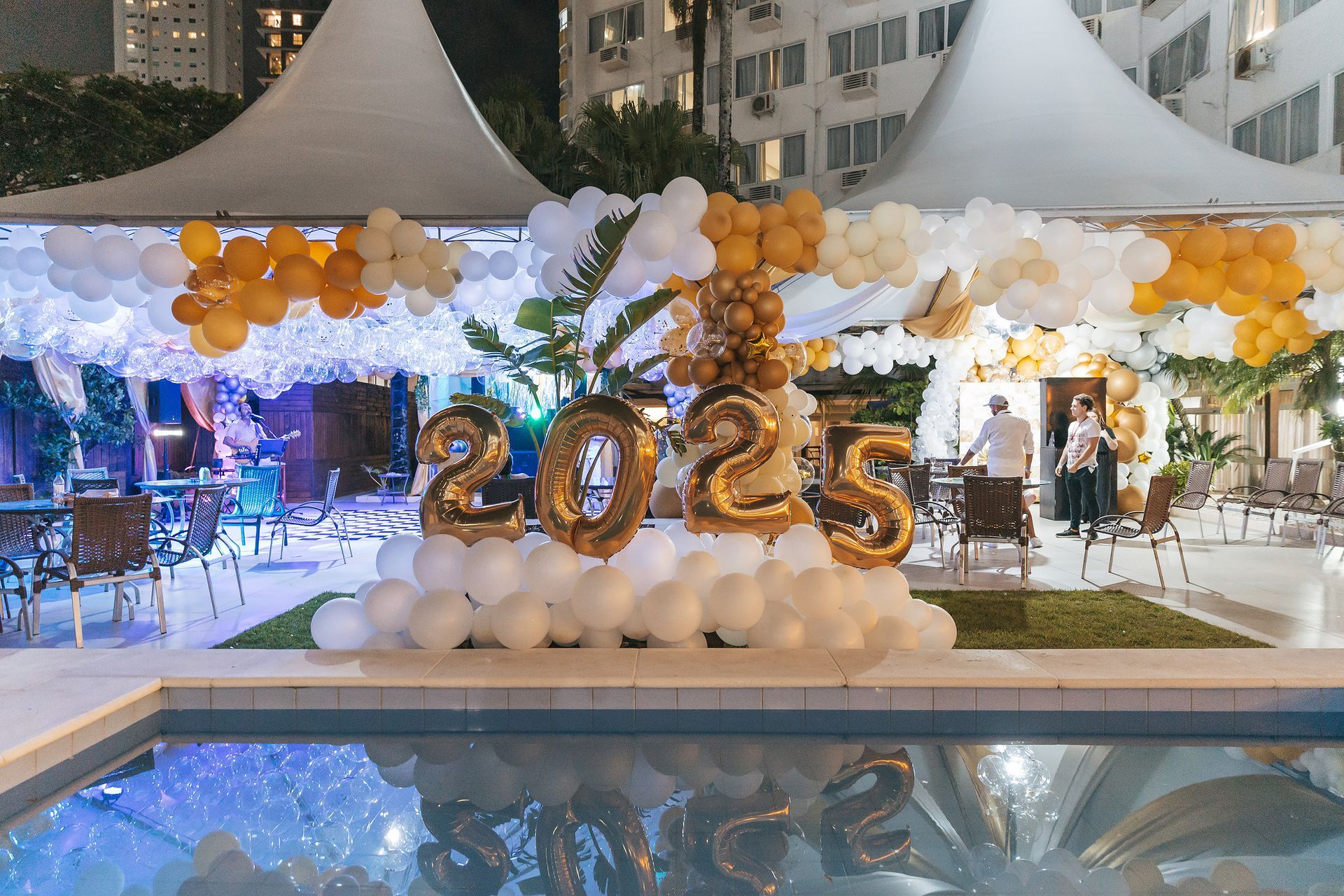 Exibição decorativa de balões dourados com o número “2025” em um evento noturno ao ar livre com decoração floral branca e dourada e reflexo na piscina.