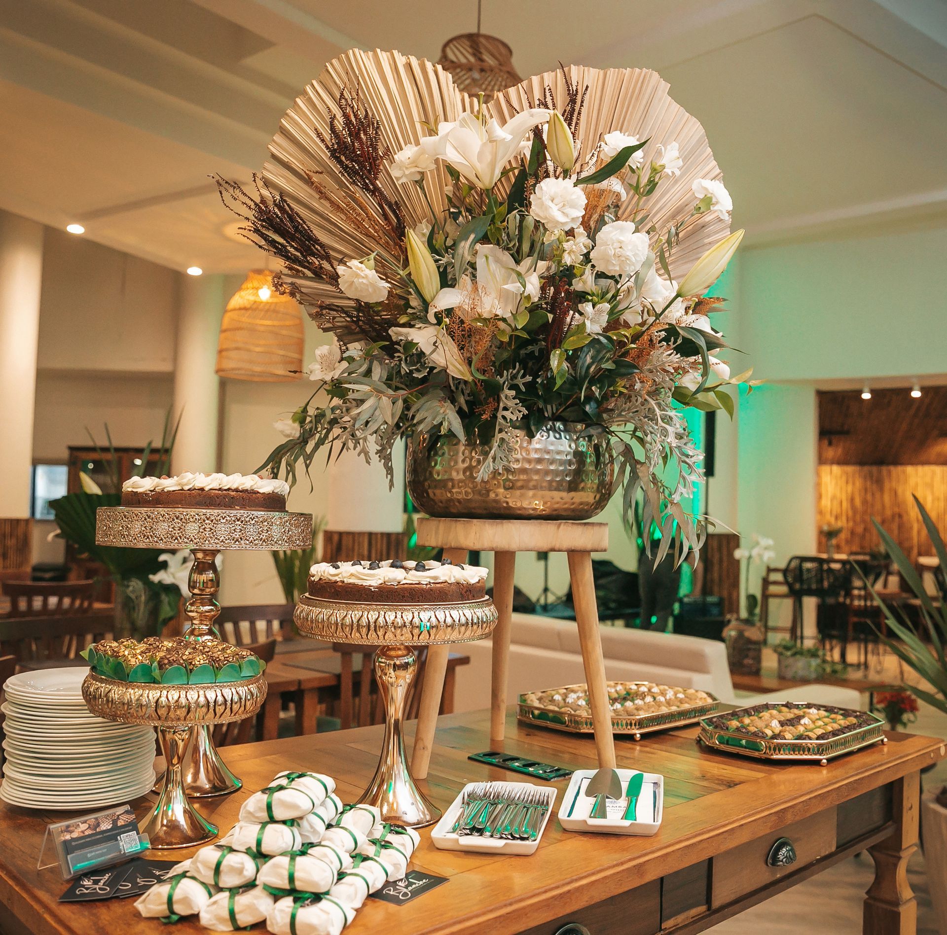 Elegante mesa de sobremesas com bolos em camadas, doces e um arranjo central alto de flores brancas em uma recepção.